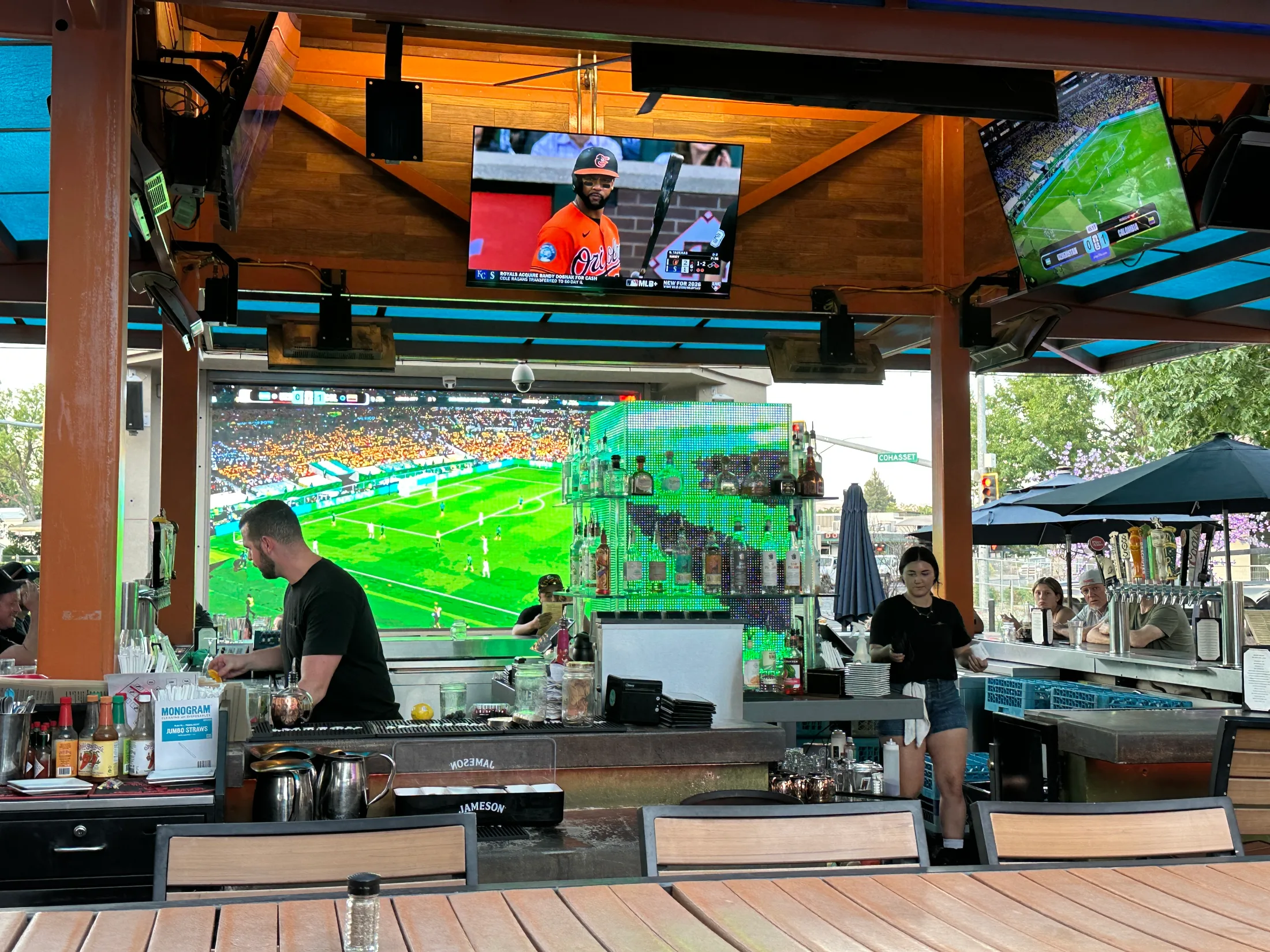 Outdoor bar at Pour House with bartender working, a large soccer match on the wall screen, and multiple TVs overhead