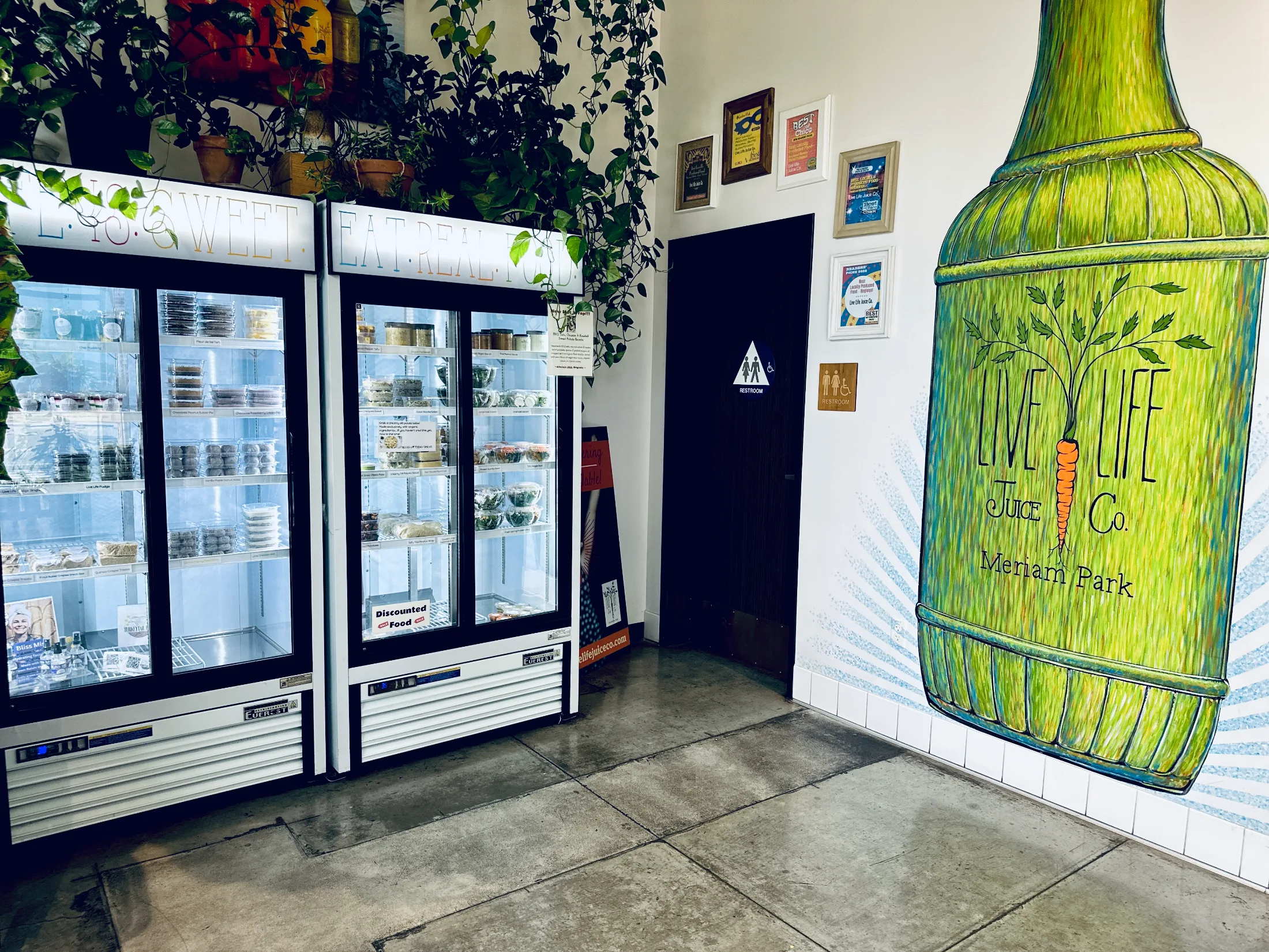 Inside Live Life Juice Co. Meriam Park showing food refrigerators, award plaques, and a large hand-painted mural of a juice bottle on the wall
