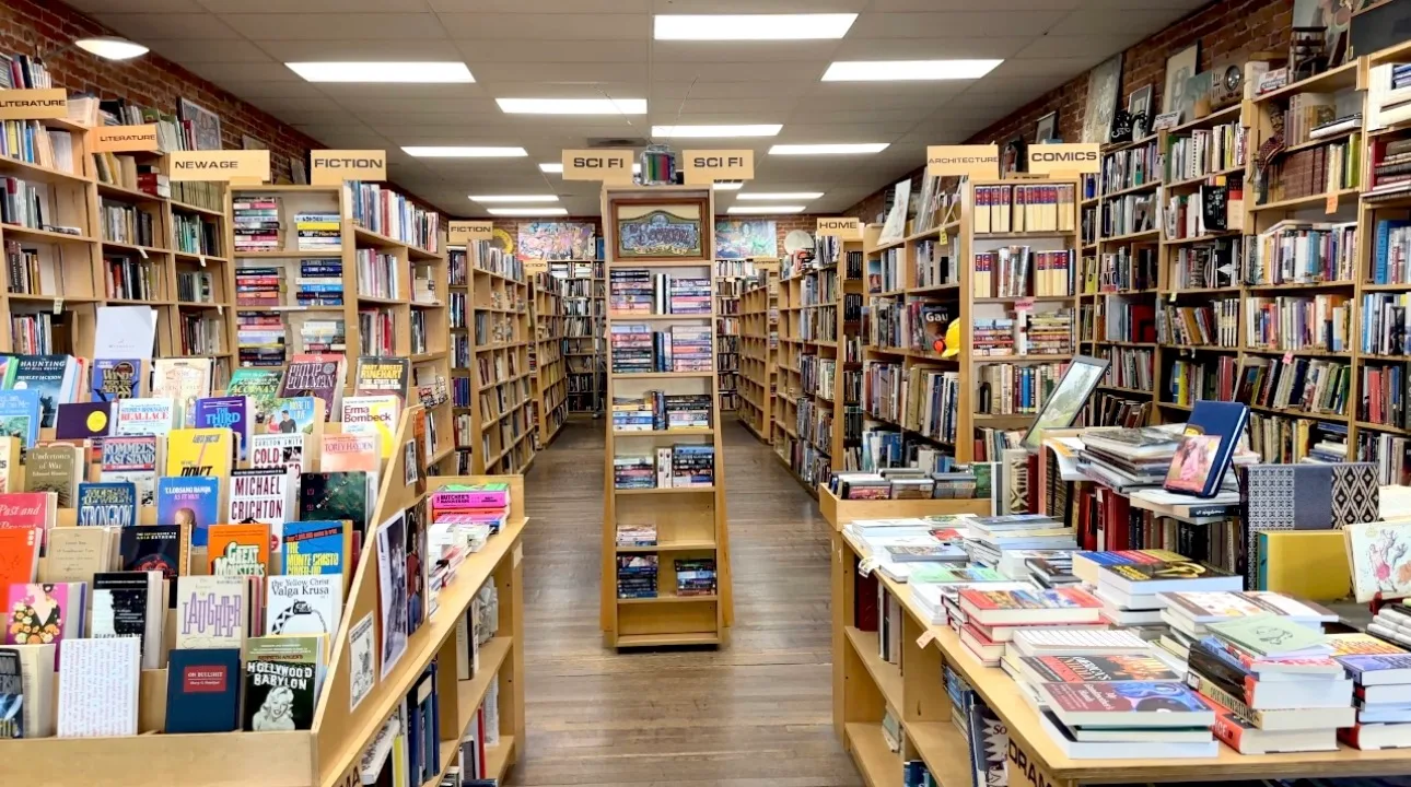 Interior of The Bookstore showing long aisles of shelved used books organized by genre including Fiction, Sci-Fi, Comics, and New Age, with exposed brick walls