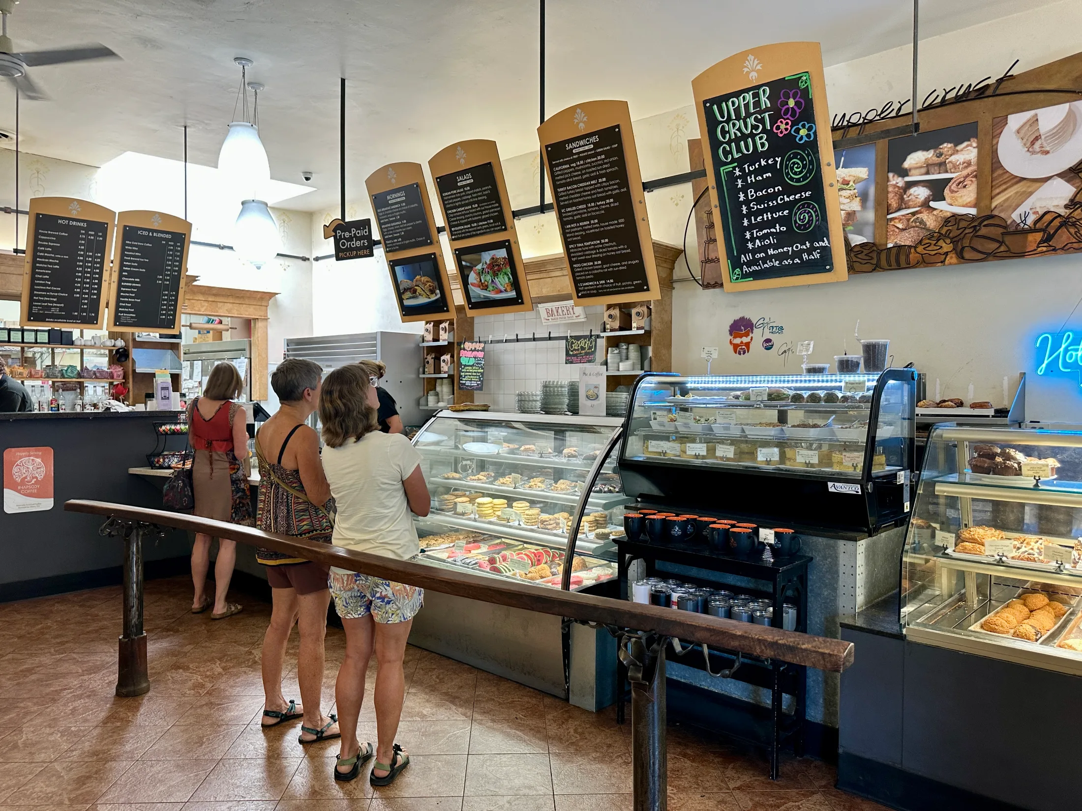 Customers studying the display cases and overhead menu boards inside Upper Crust Bakery