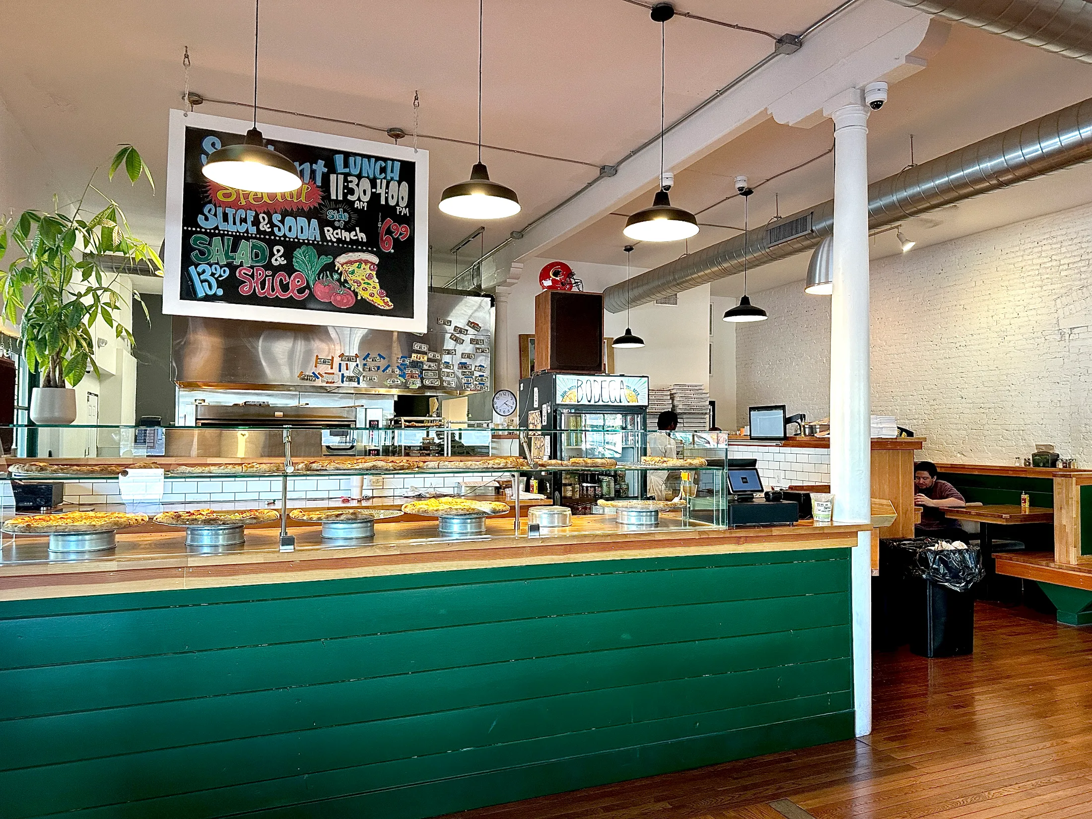 Bright interior of Bodega Slice Shop showing the full counter display, hanging specials chalkboard, and green-paneled booths