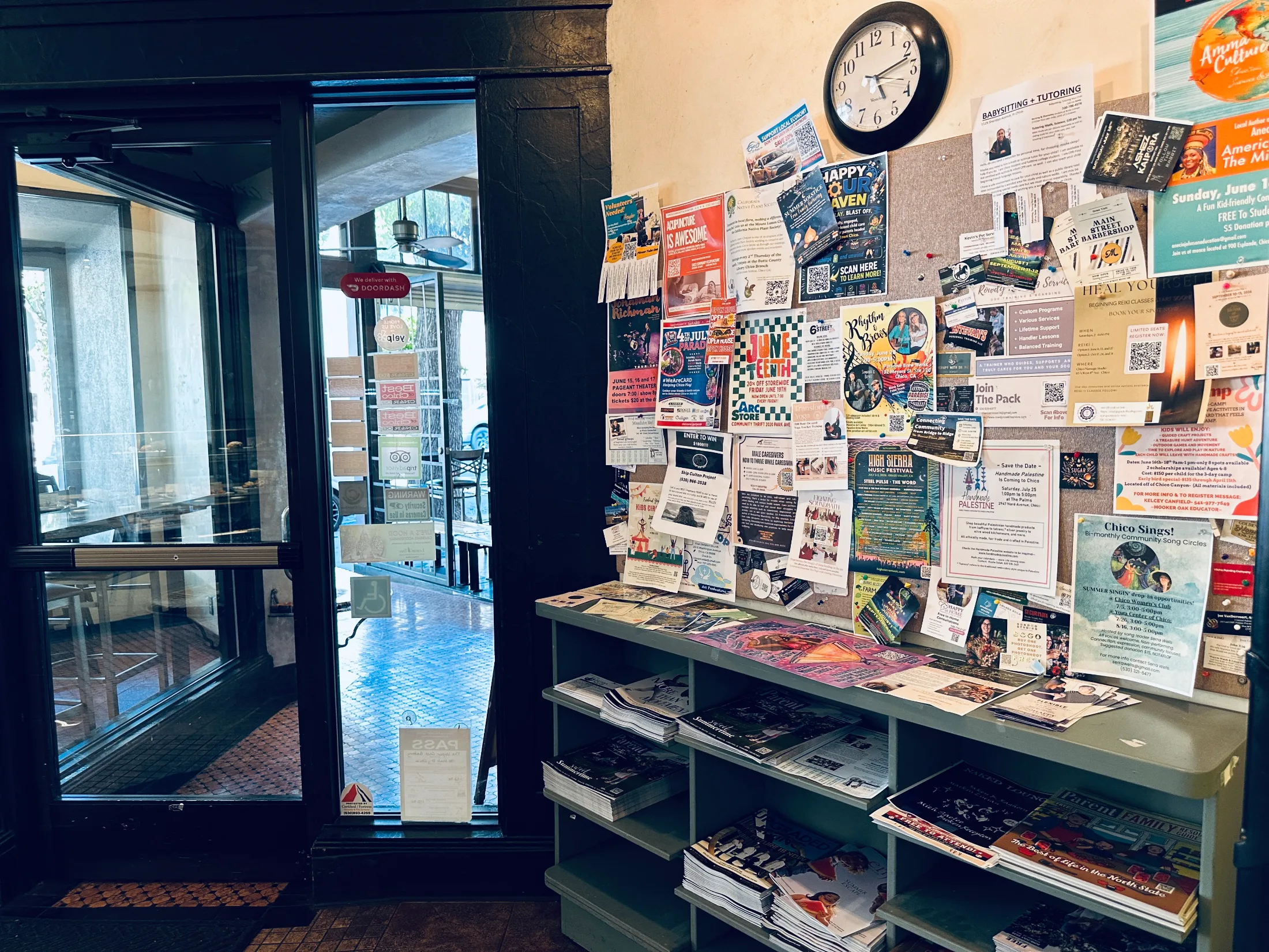 Community corkboard near the entrance of Upper Crust Bakery with flyers for High Sierra Music Festival, Chico Sings, local services, and free magazines below