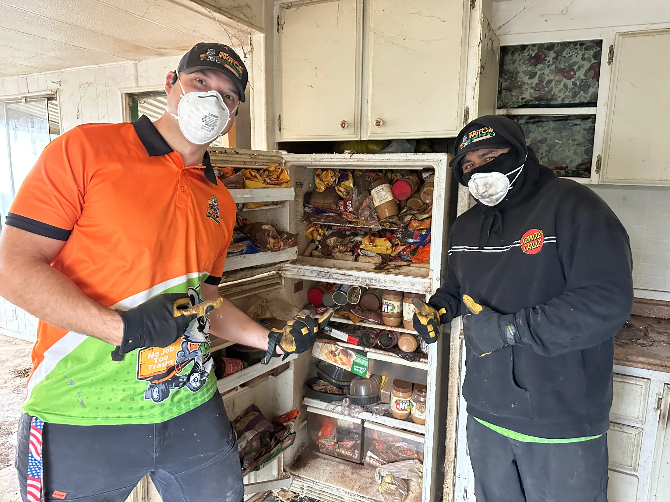 Two NorCal Trash Pandas crew members in protective gloves and masks clearing out a severely cluttered refrigerator inside a hoarder house