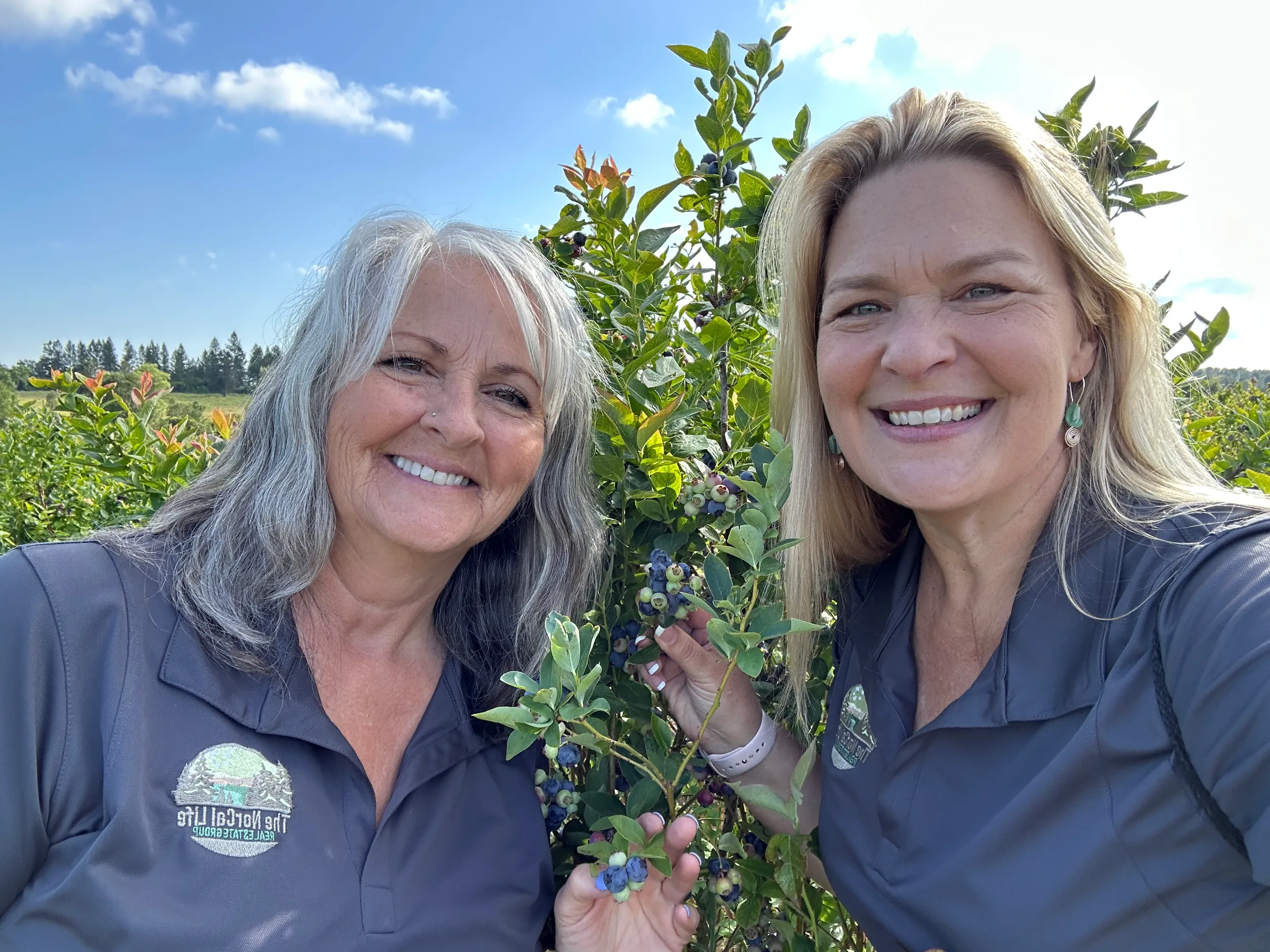 Two women smiling together in the blueberry rows on a sunny morning
