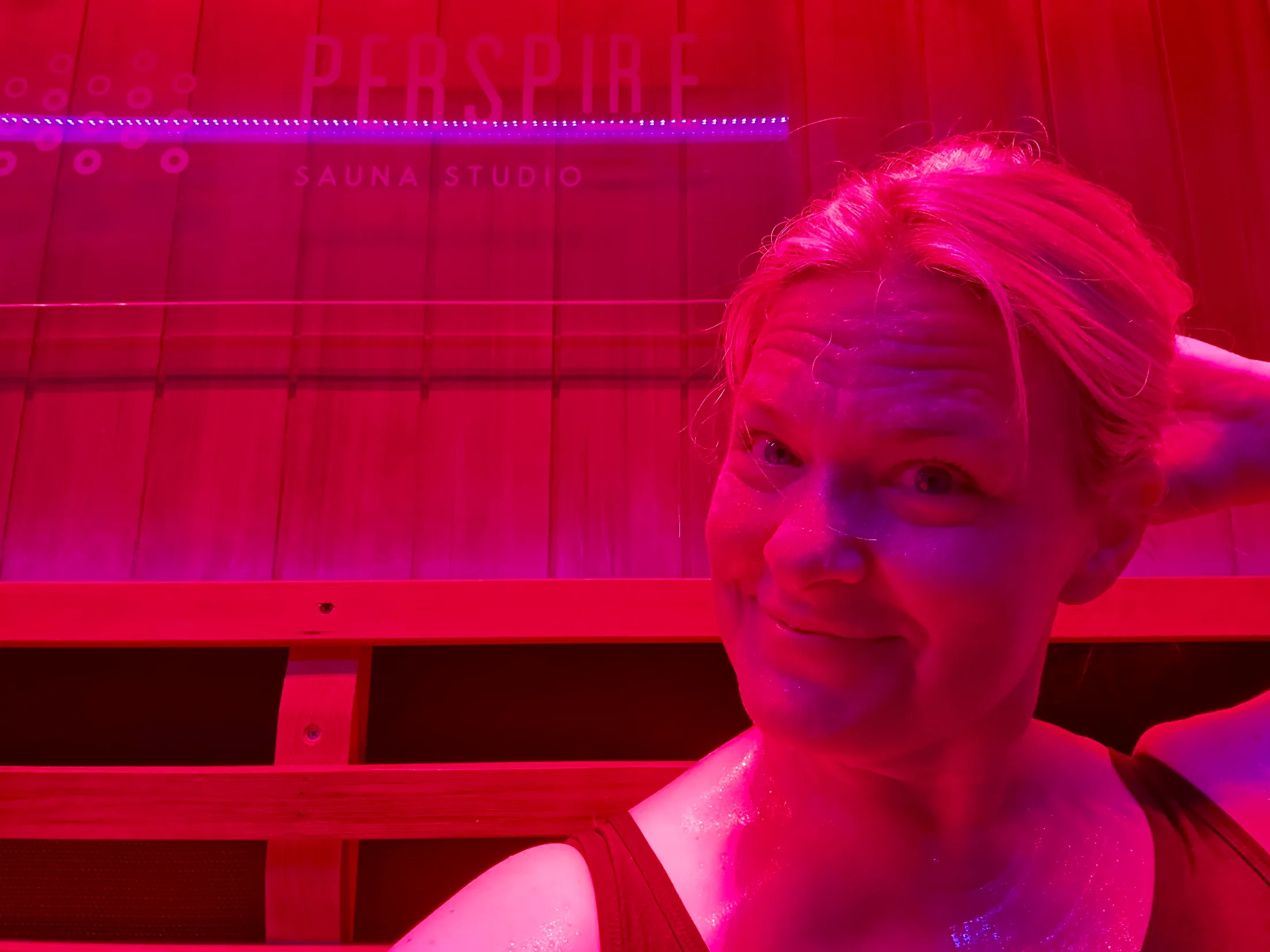 Woman smiling inside the red-lit infrared sauna suite at Perspire Sauna Studio