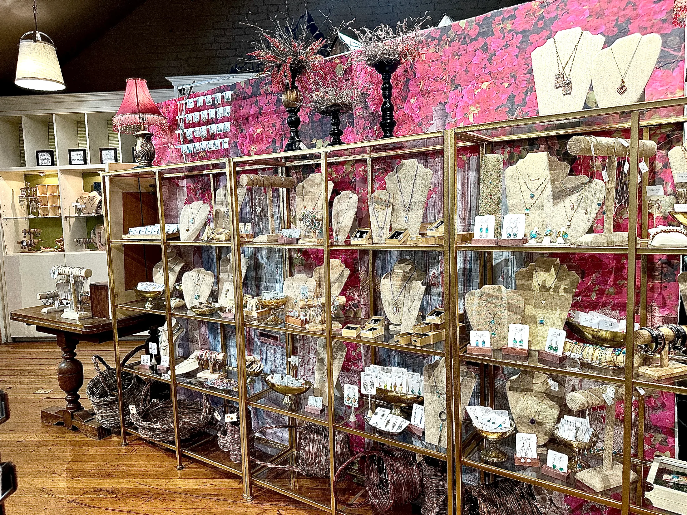 Gold-framed glass jewelry display cases at Made in Chico showing necklaces, earrings, and pendants from local makers, against a floral backdrop