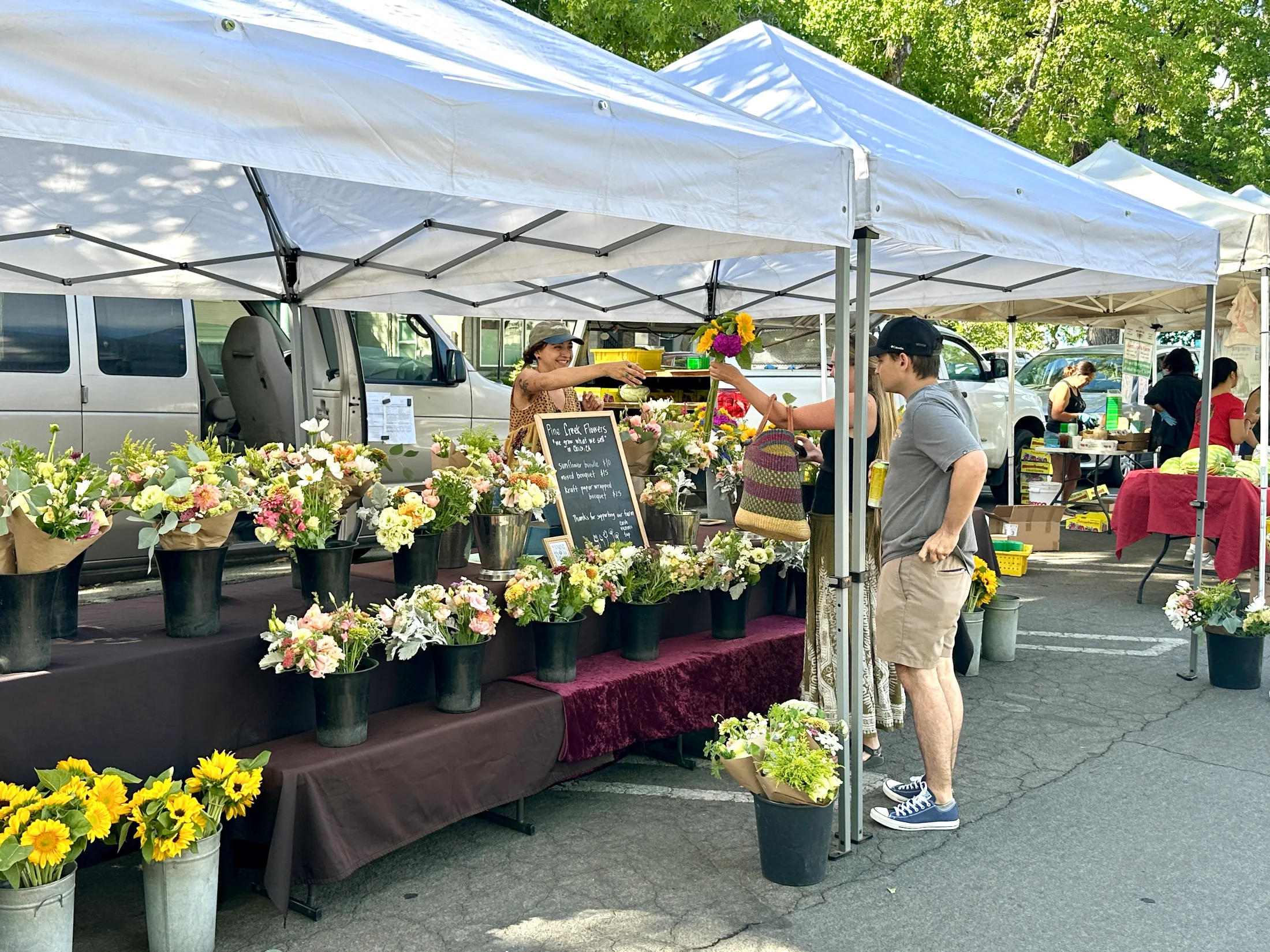 Flower vendor handing a bouquet to a customer at a farmers market stall with buckets of blooms