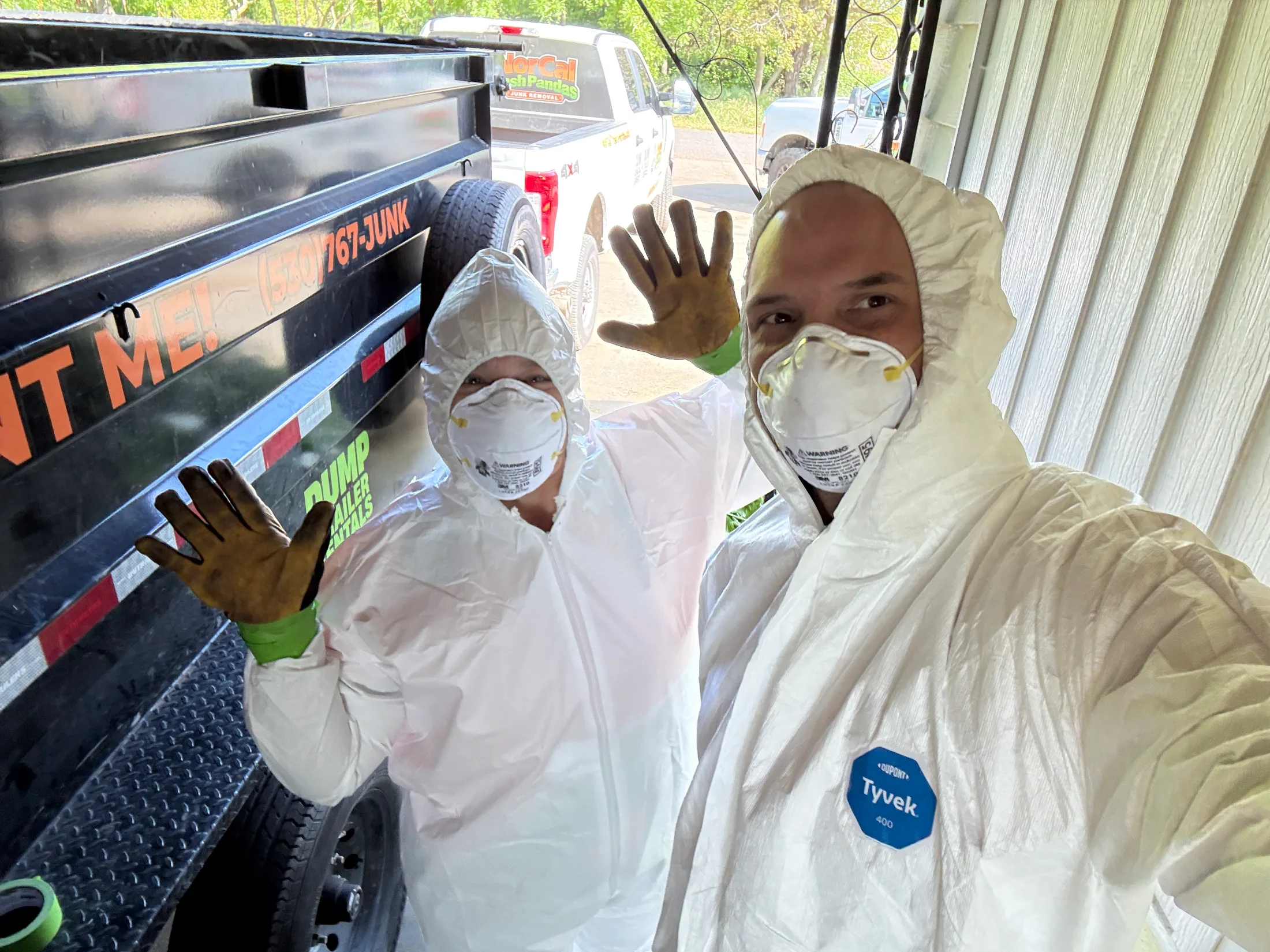Two NorCal Trash Pandas crew members in full Tyvek hazmat suits waving outside a home before a hazmat cleanout job