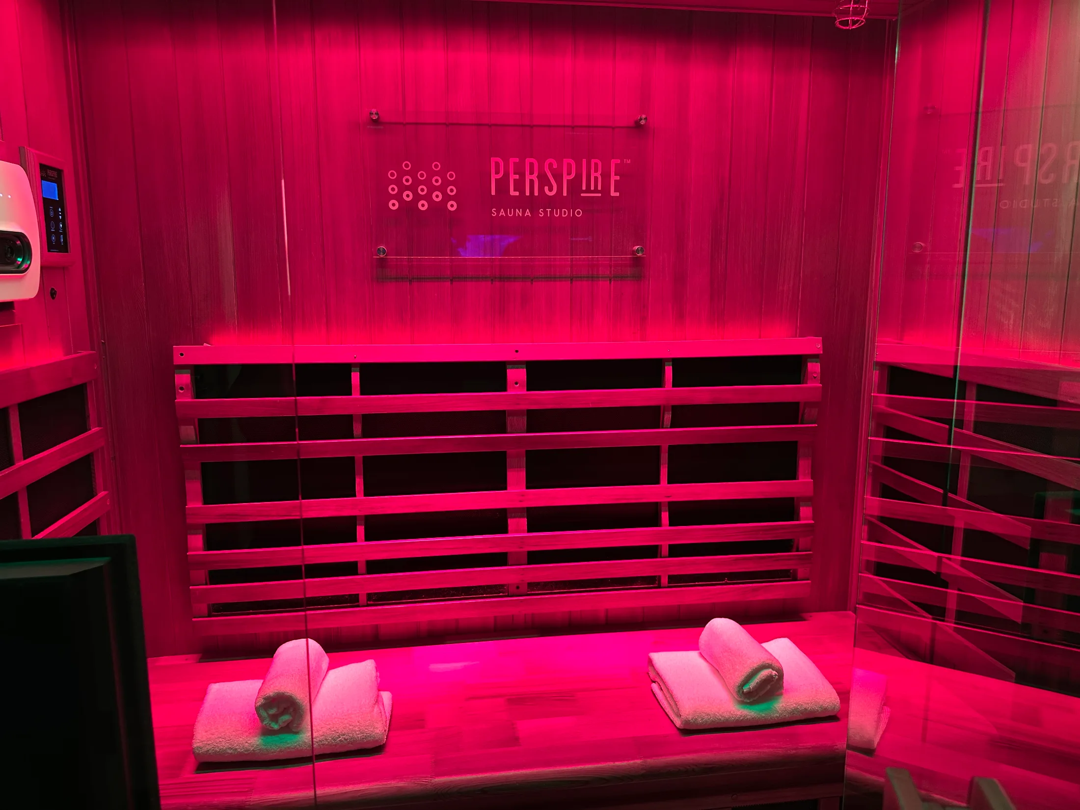 Private infrared sauna suite at Perspire Sauna Studio bathed in red light, two rolled towels on the bench