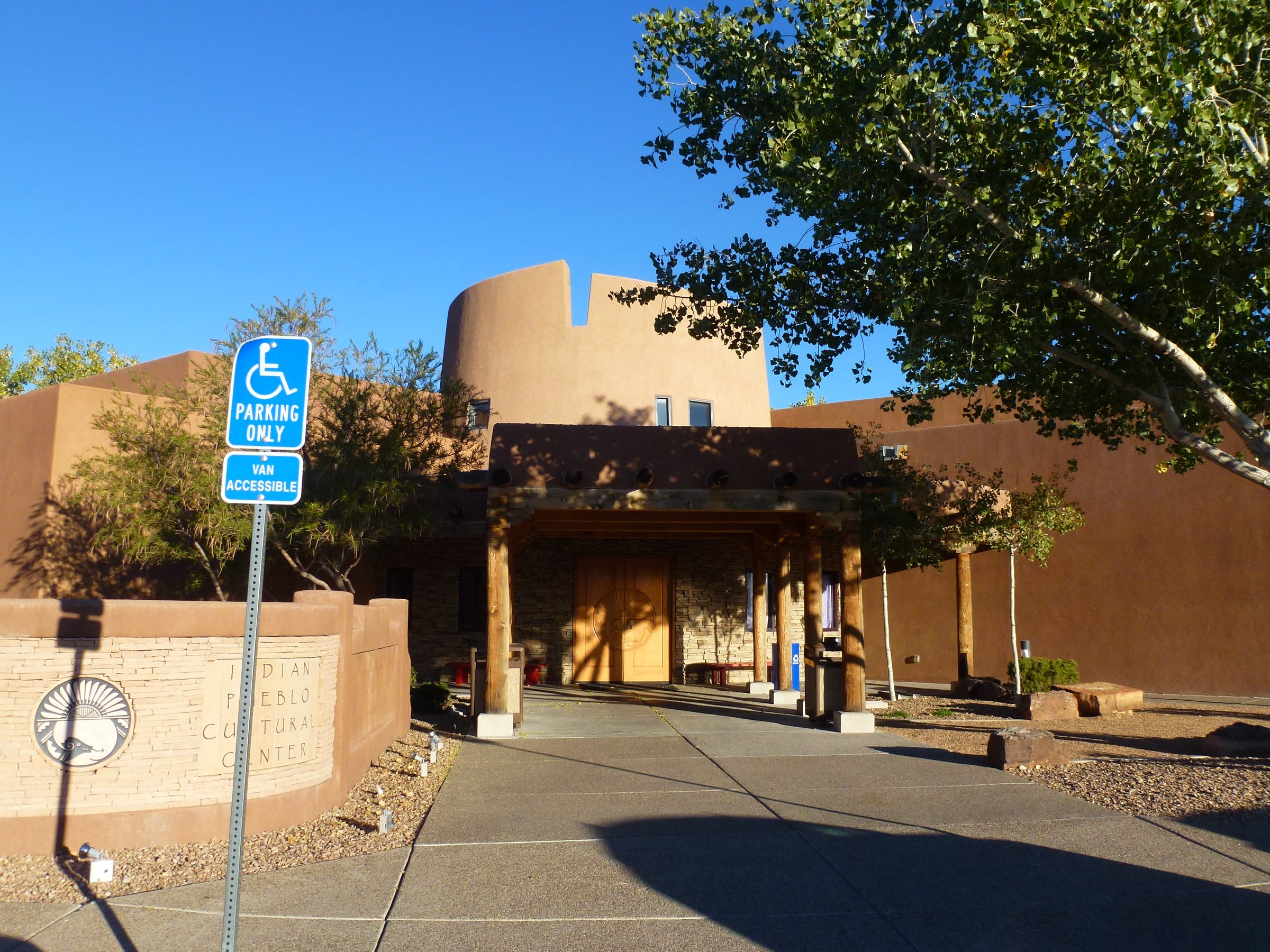The entrance of the Indian Pueblo Cultural Center, a tan building with a wooden portal and trees