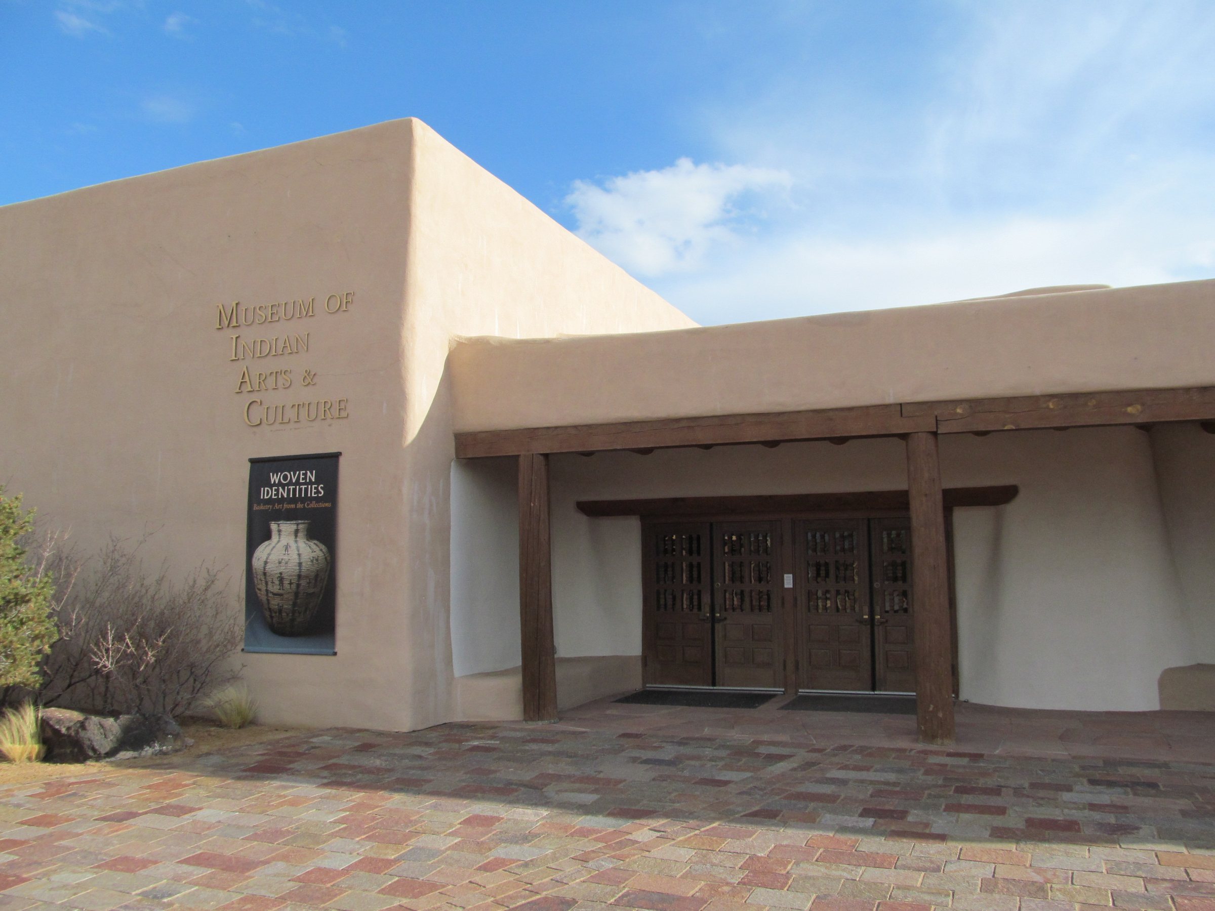The Museum of Indian Arts and Culture entrance, a tan adobe-style building with wooden posts and a patterned plaza
