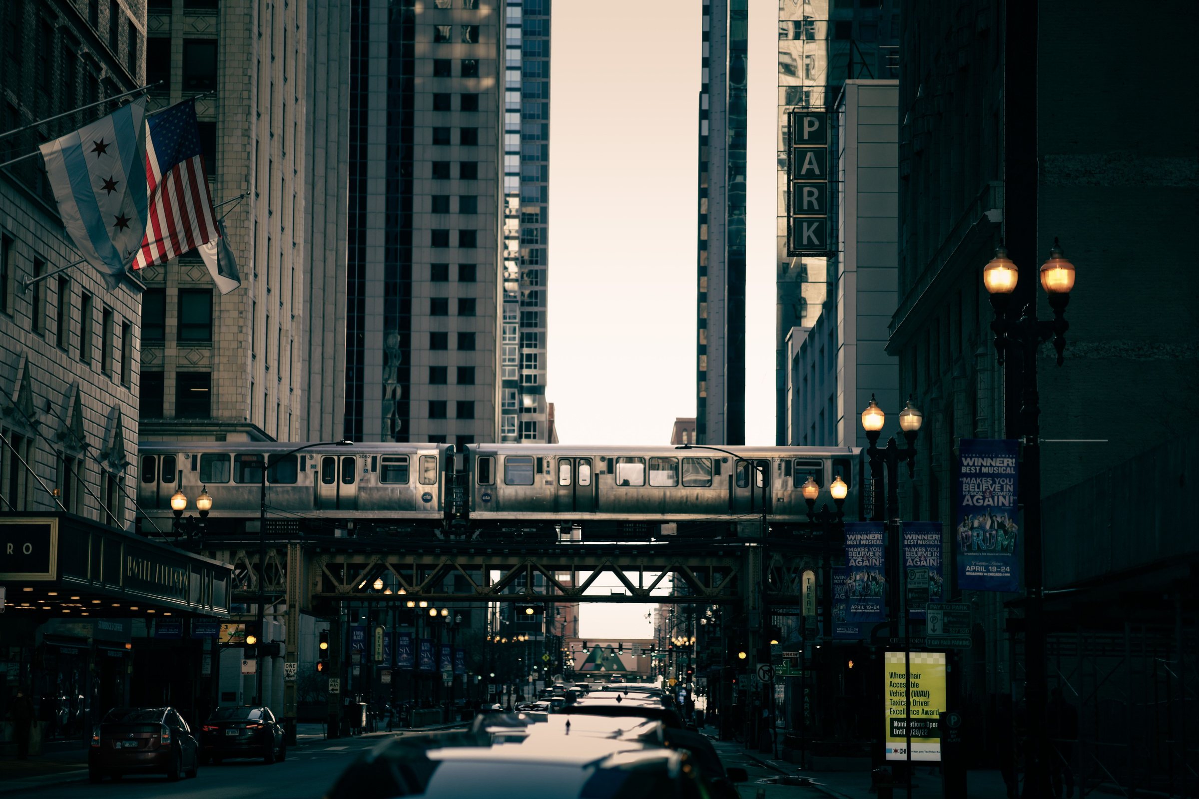 A silver elevated train on a steel rail bridge between tall buildings, with a Chicago city flag and an American flag hanging from a building on the left