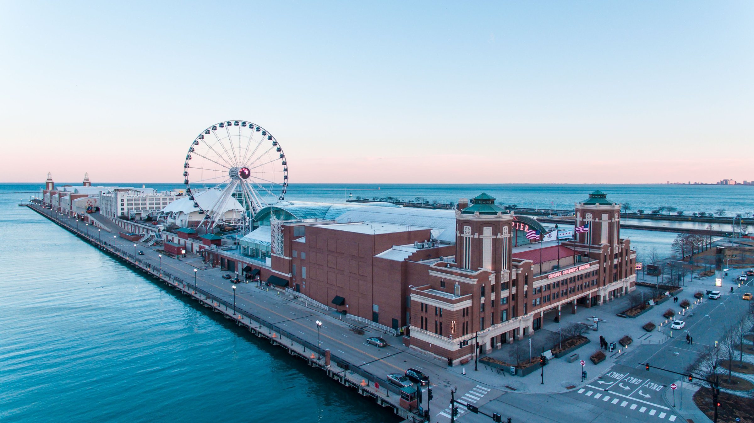 Aerial view of a long pier reaching into blue water under a pink and blue sky, with a large Ferris wheel on the pier and a red-brick building with towers at its land end