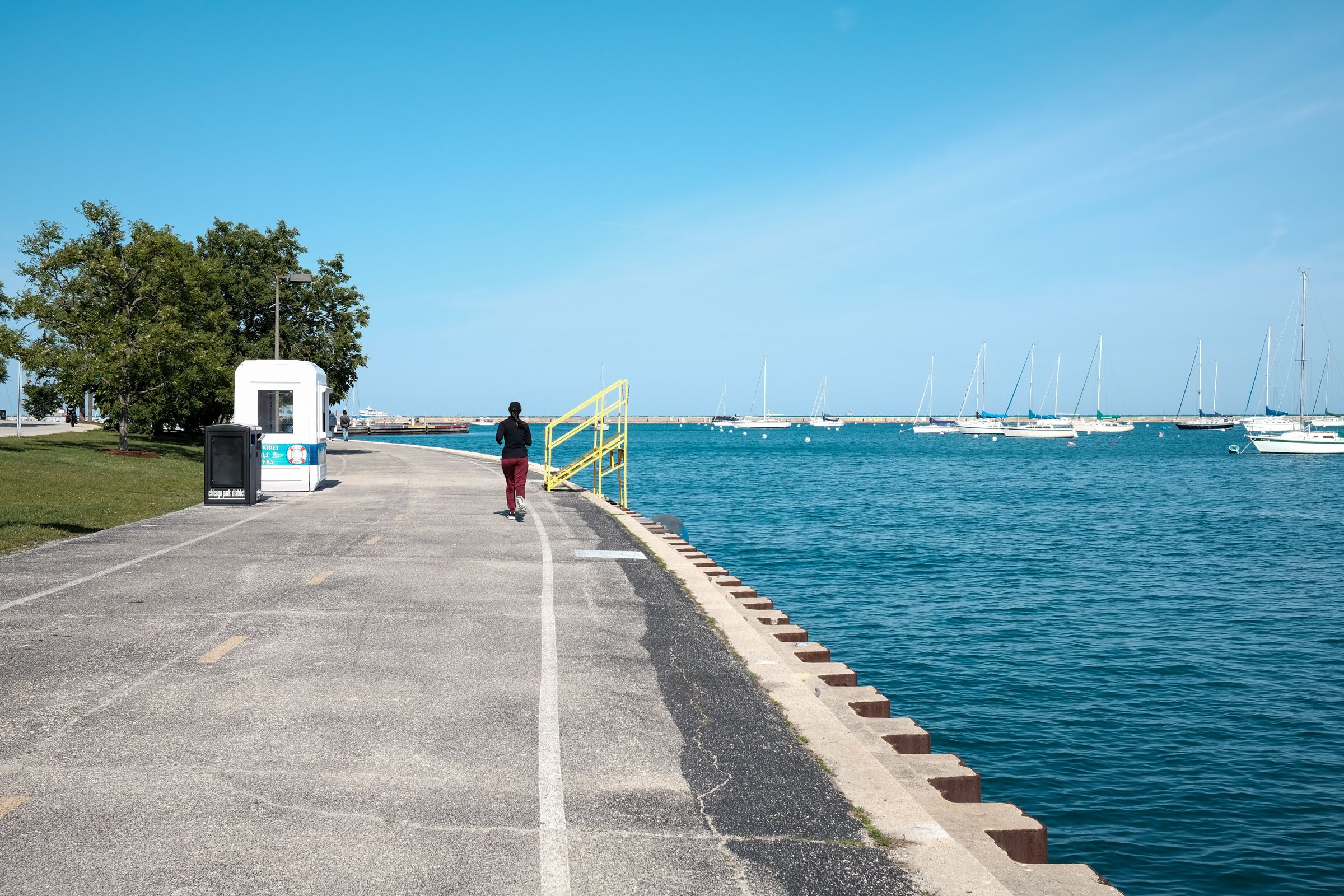 A wide paved path with a painted white center line beside a concrete seawall and blue water, with one person running on the path