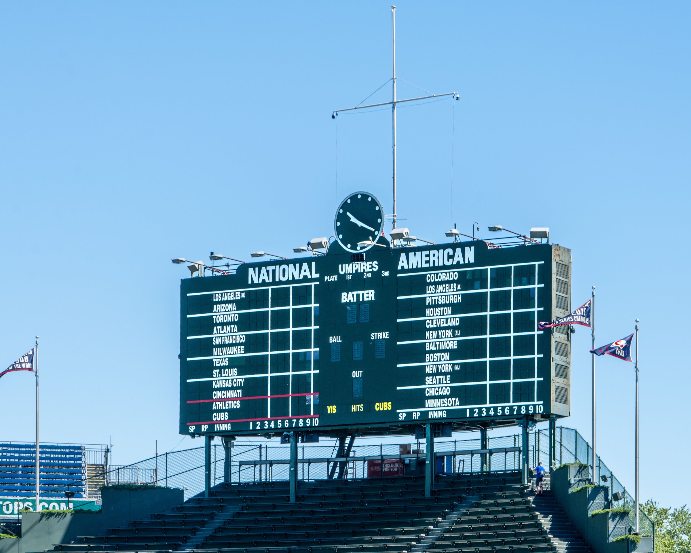 A large dark green manual scoreboard with white lettering reading NATIONAL and AMERICAN and a round clock on top, above rows of empty green bleacher seats