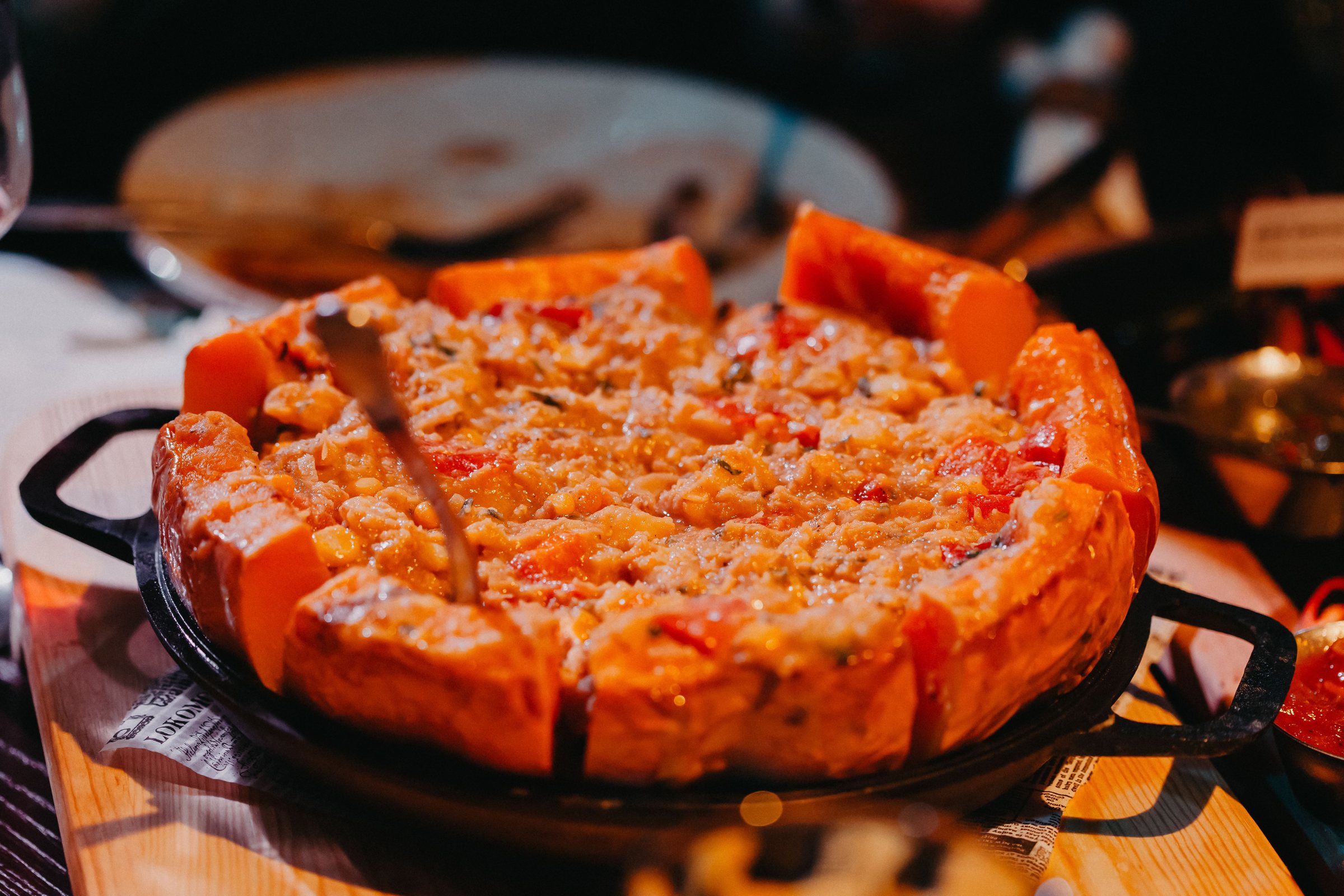 A deep, round pizza in a black cast-iron pan on a wooden board, with a tall golden crust edge and a top of cheese and chopped red tomato