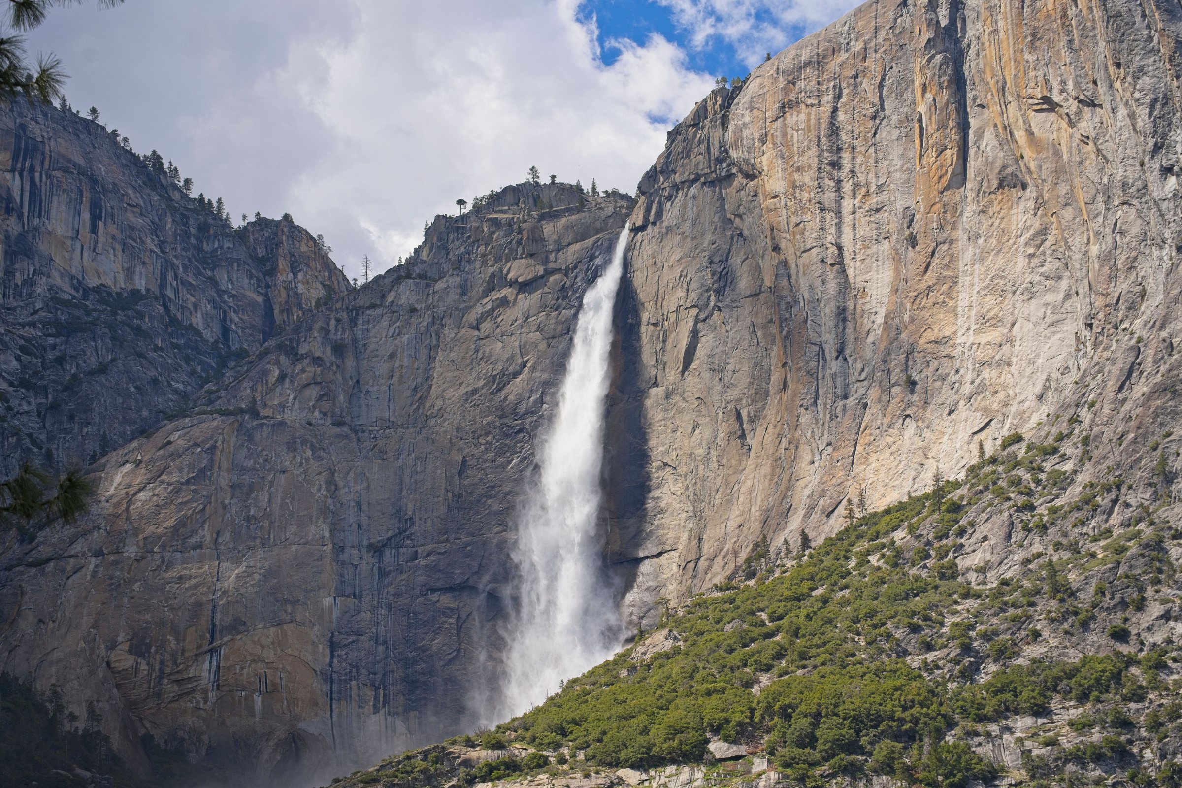 A tall white waterfall pours down a gap in sheer gray granite cliffs, with green trees at the base