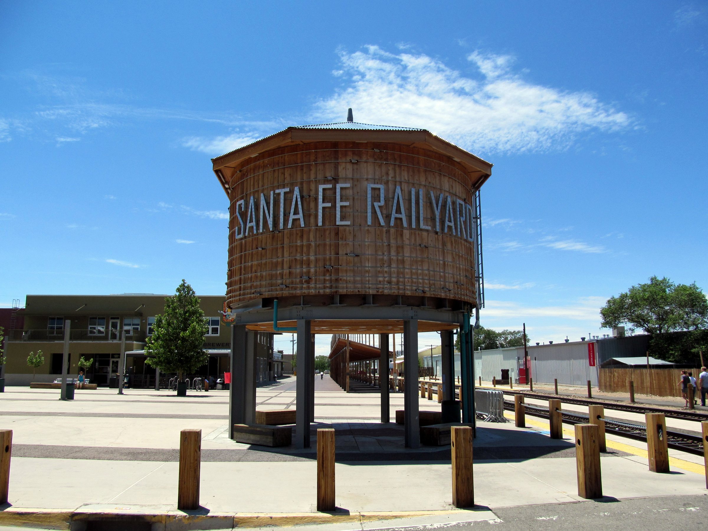 A round wooden water tower painted with the words Santa Fe Railyard, standing on metal legs on a plaza under a blue sky