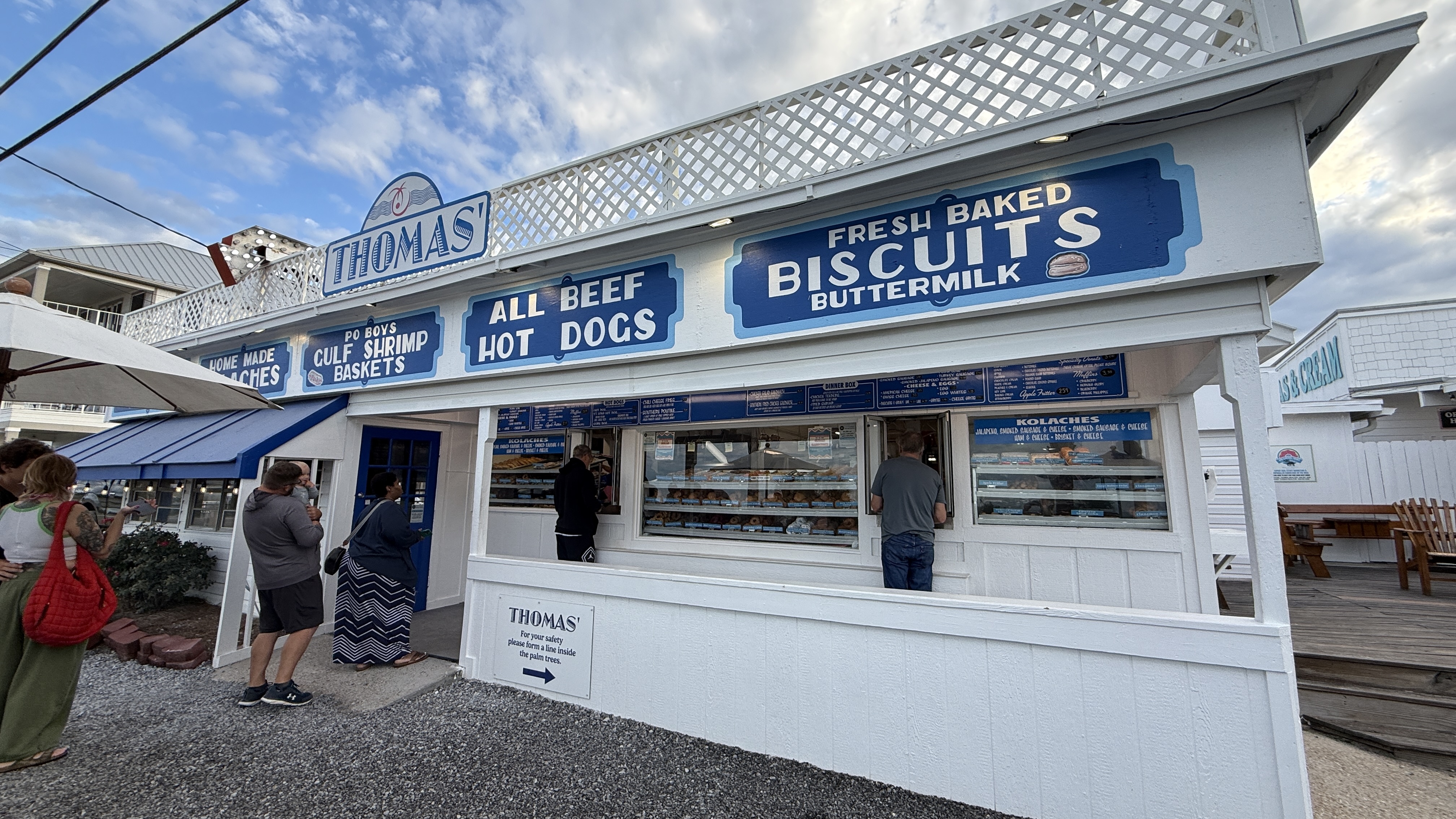 Thomas' Donuts walk-up counter in Panama City Beach, blue and white signage advertising fresh baked buttermilk biscuits, gulf shrimp baskets, kolaches, and all-beef hot dogs, with customers ordering at the windows