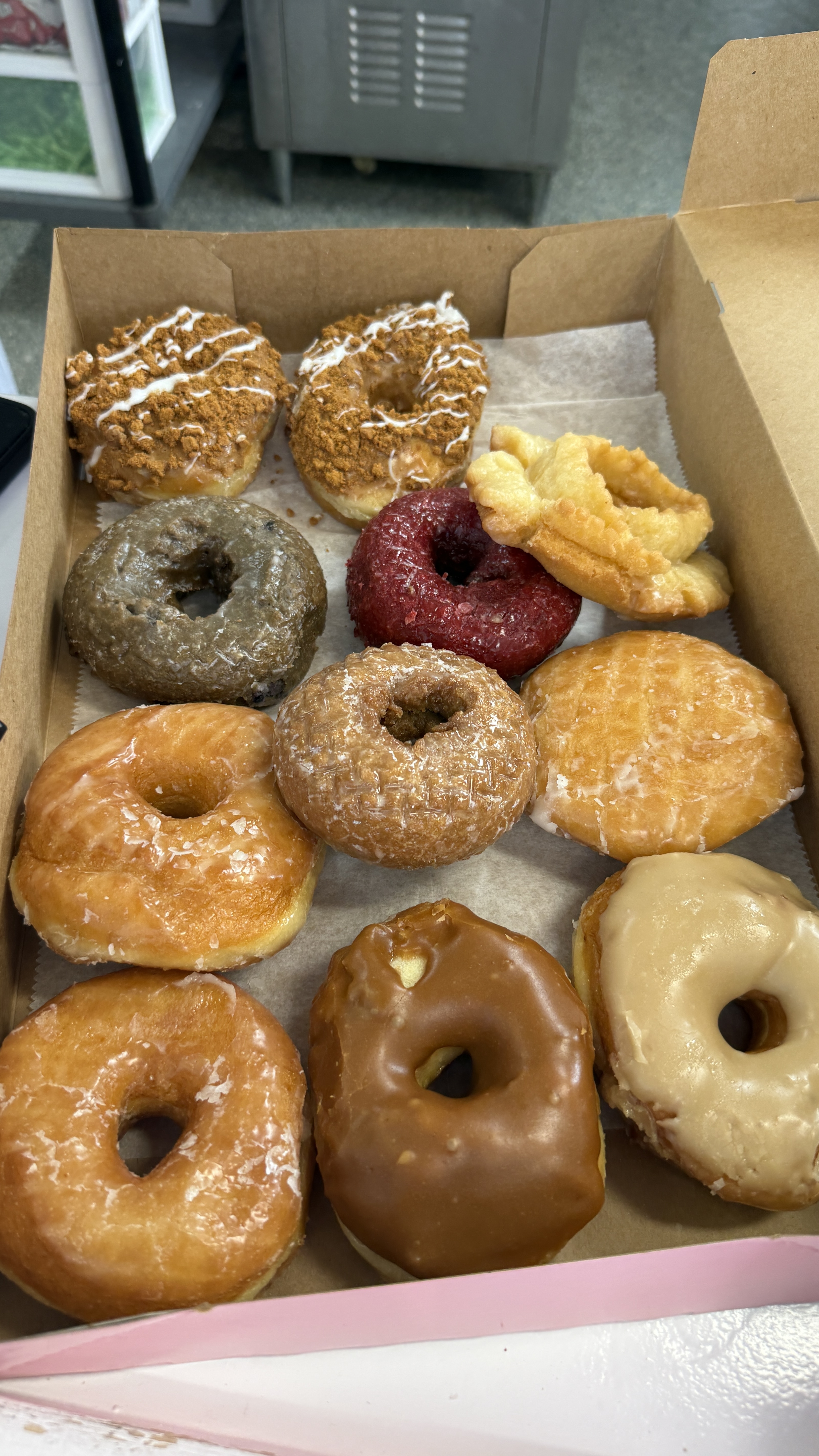 Open box of a dozen Thomas' Donuts varieties including glazed, chocolate-dipped, crumb-topped, a red-glazed donut, a cruller, and specialty rings