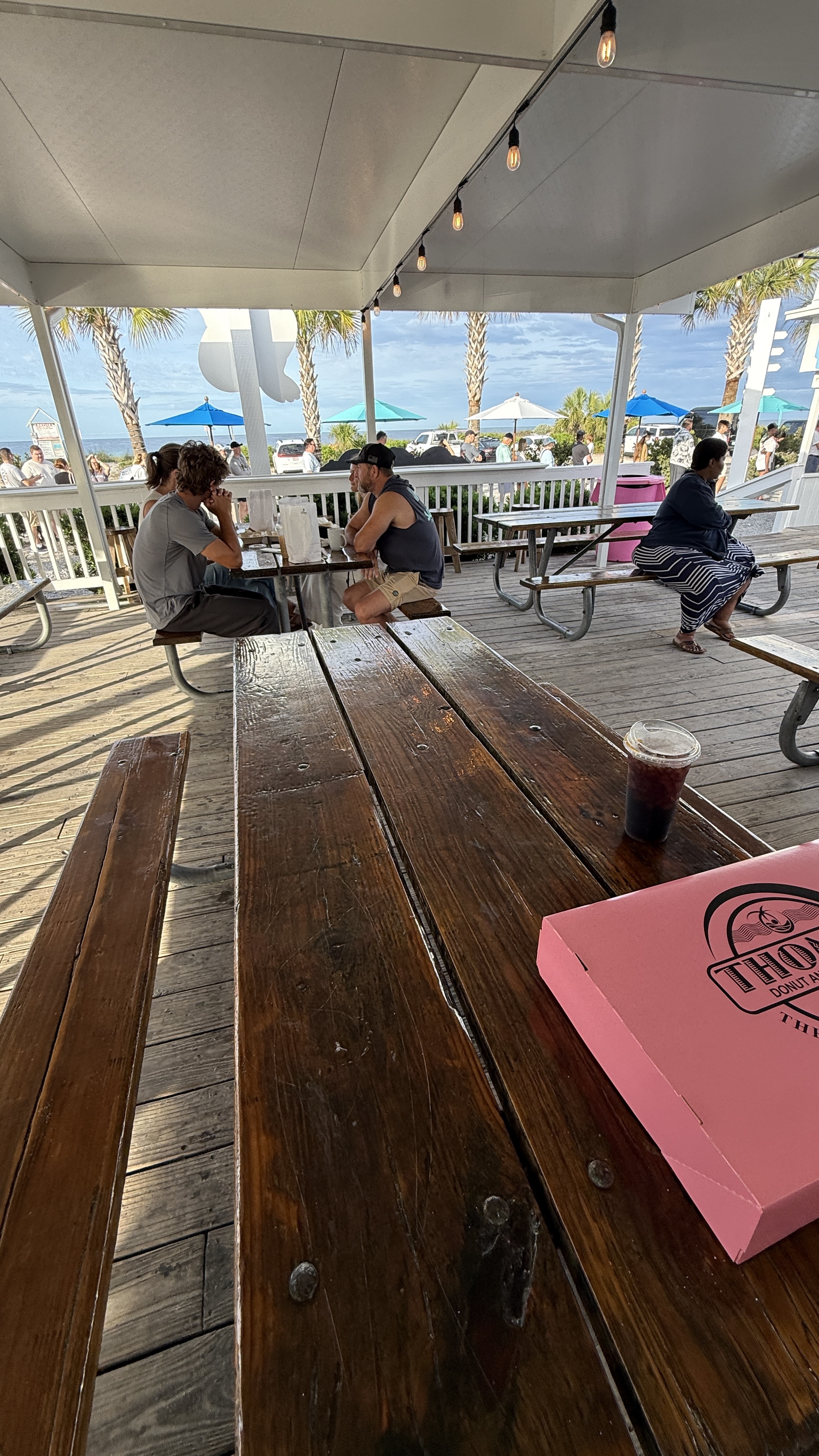 Covered outdoor deck at Thomas' Donuts with picnic tables, string lights, palm trees, and a view of the Gulf of Mexico in the background