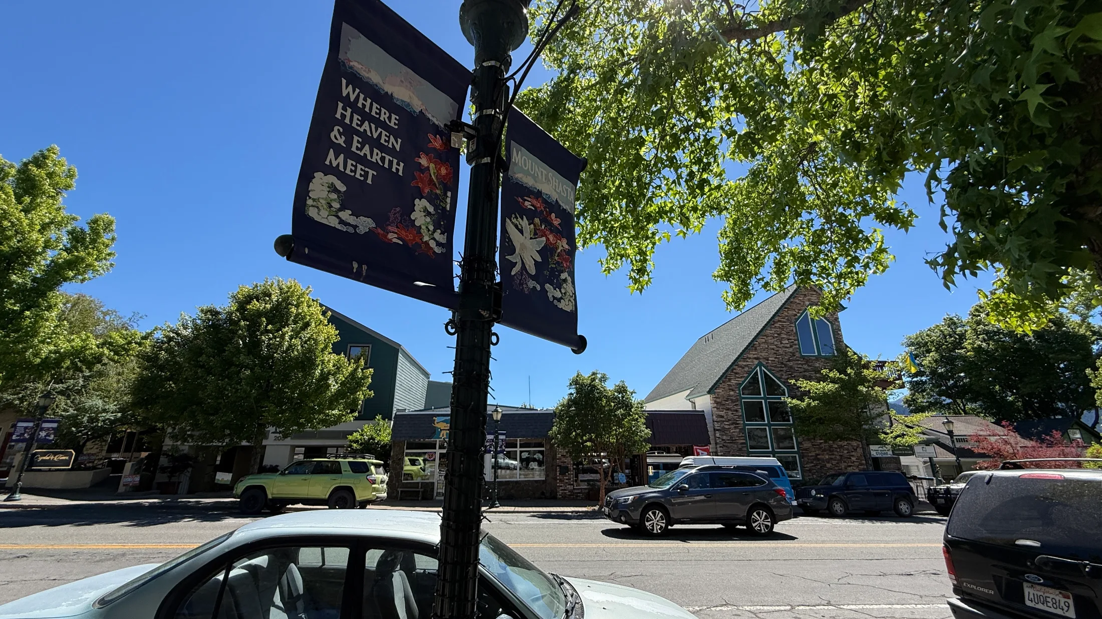 Downtown Mount Shasta lamp post banners reading 'Where Heaven and Earth Meet' on a sunny summer afternoon with green trees and local storefronts behind