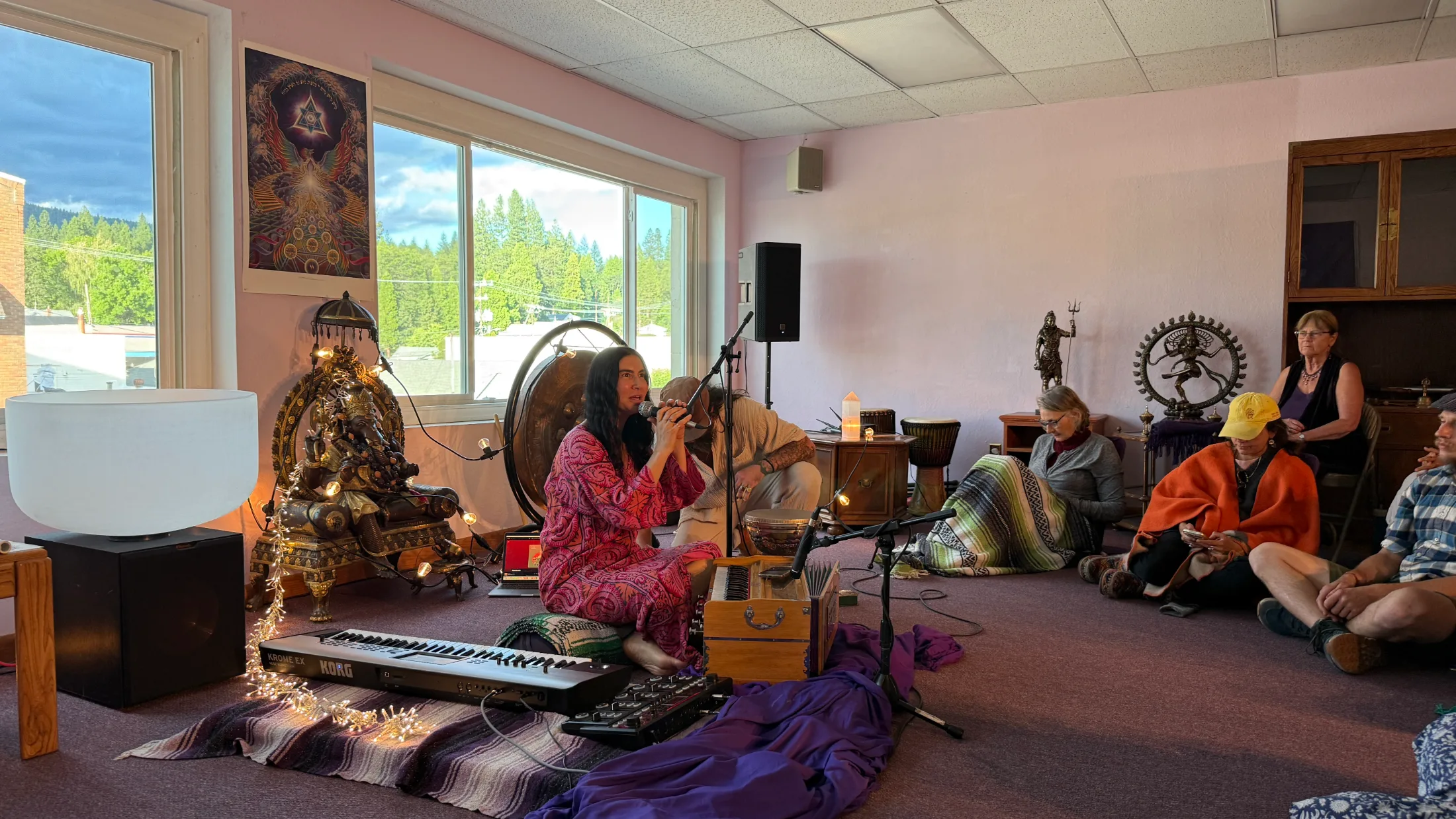Simrit Kaur holding a microphone with both hands, singing deeply, participants seated behind her wrapped in blankets