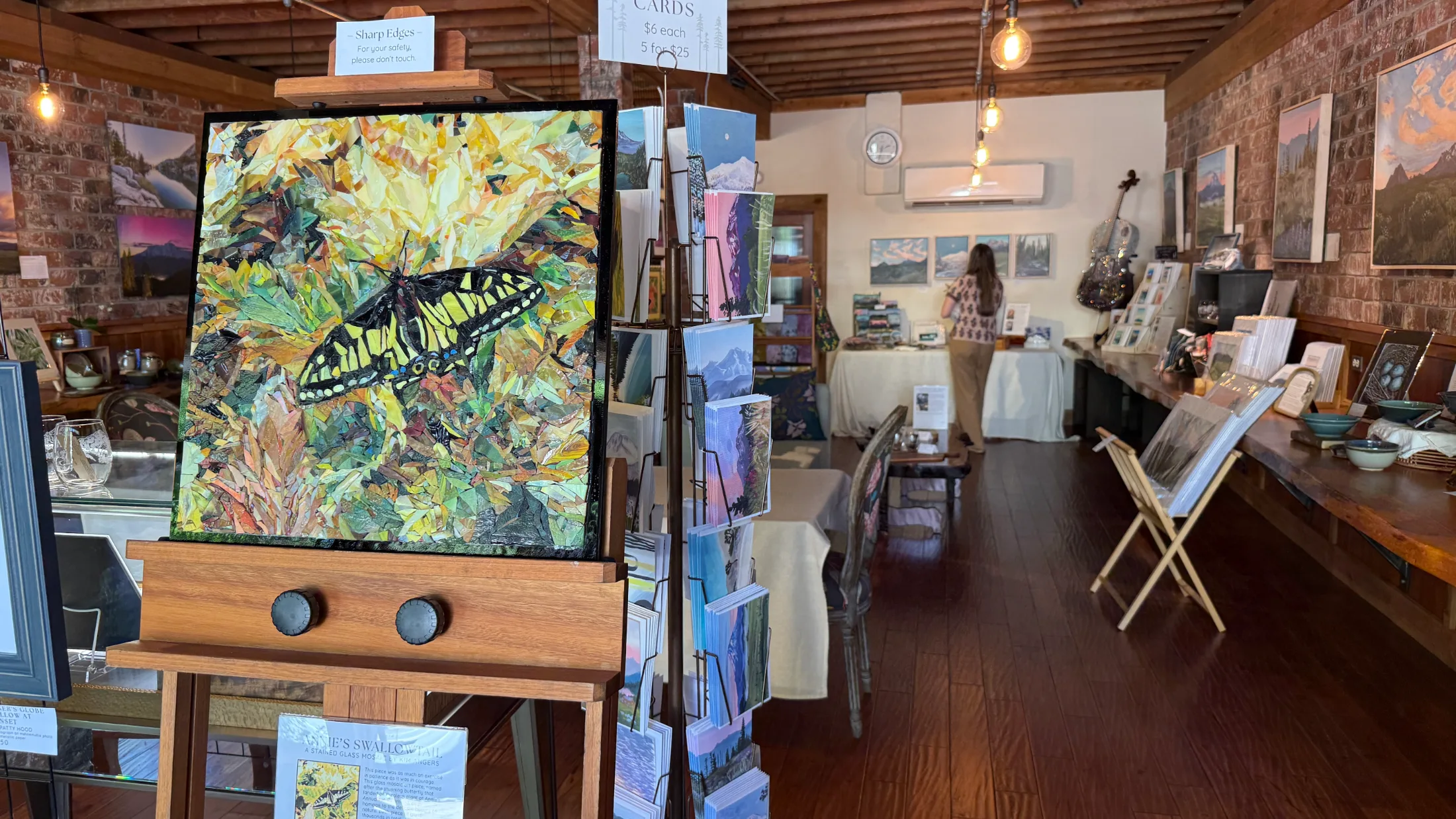 Stained glass mosaic butterfly painting on easel in the foreground with art cards and mountain landscape paintings visible throughout the gallery