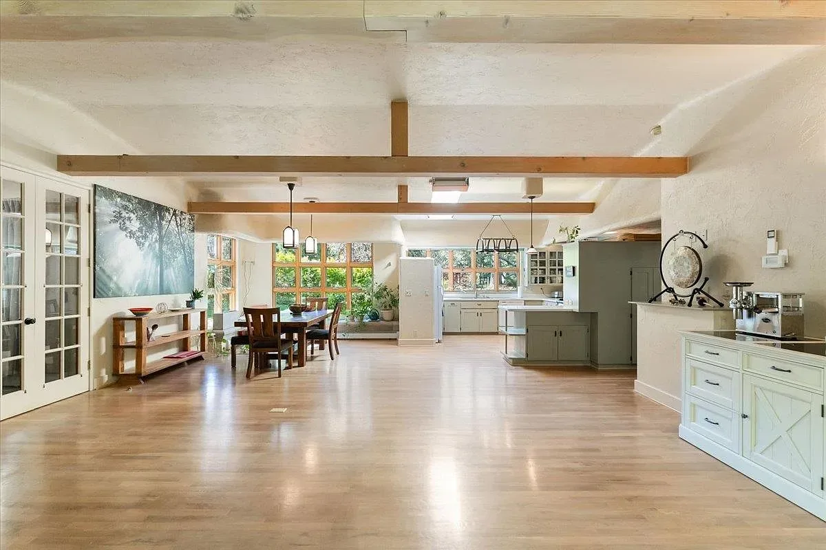 Open-plan kitchen and dining room with sage-green cabinetry, wood beams, and grid windows overlooking greenery