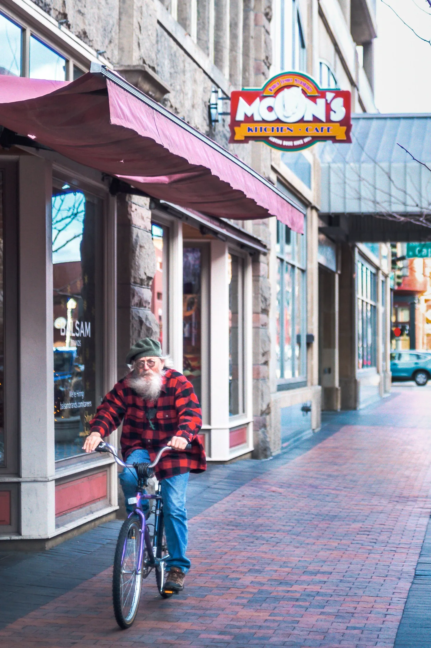 An older bearded man in a red flannel rides a purple cruiser bicycle along a brick sidewalk past storefronts