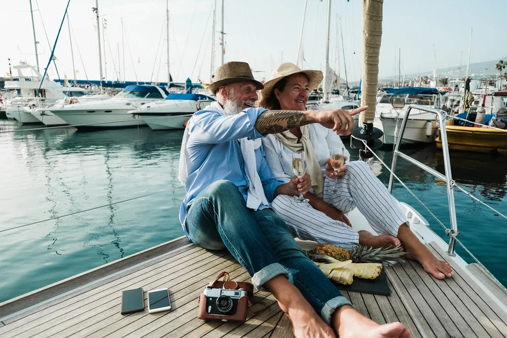 Two people relaxing on a sailboat in a sun-bright marina, holding glasses of wine, pointing toward the water