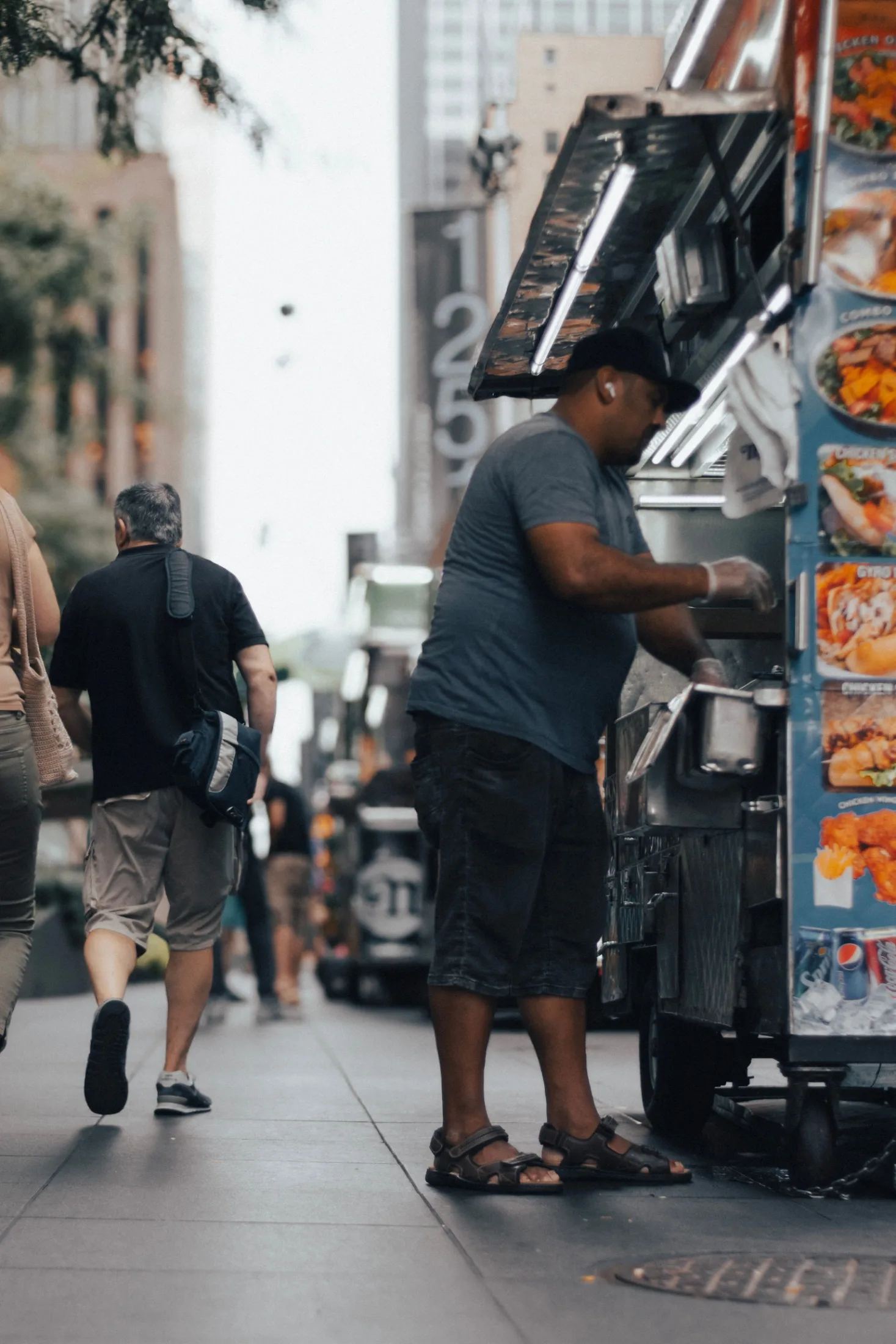 A food cart vendor working the grill on a midtown sidewalk, pedestrians walking past in the background, city buildings rising behind him