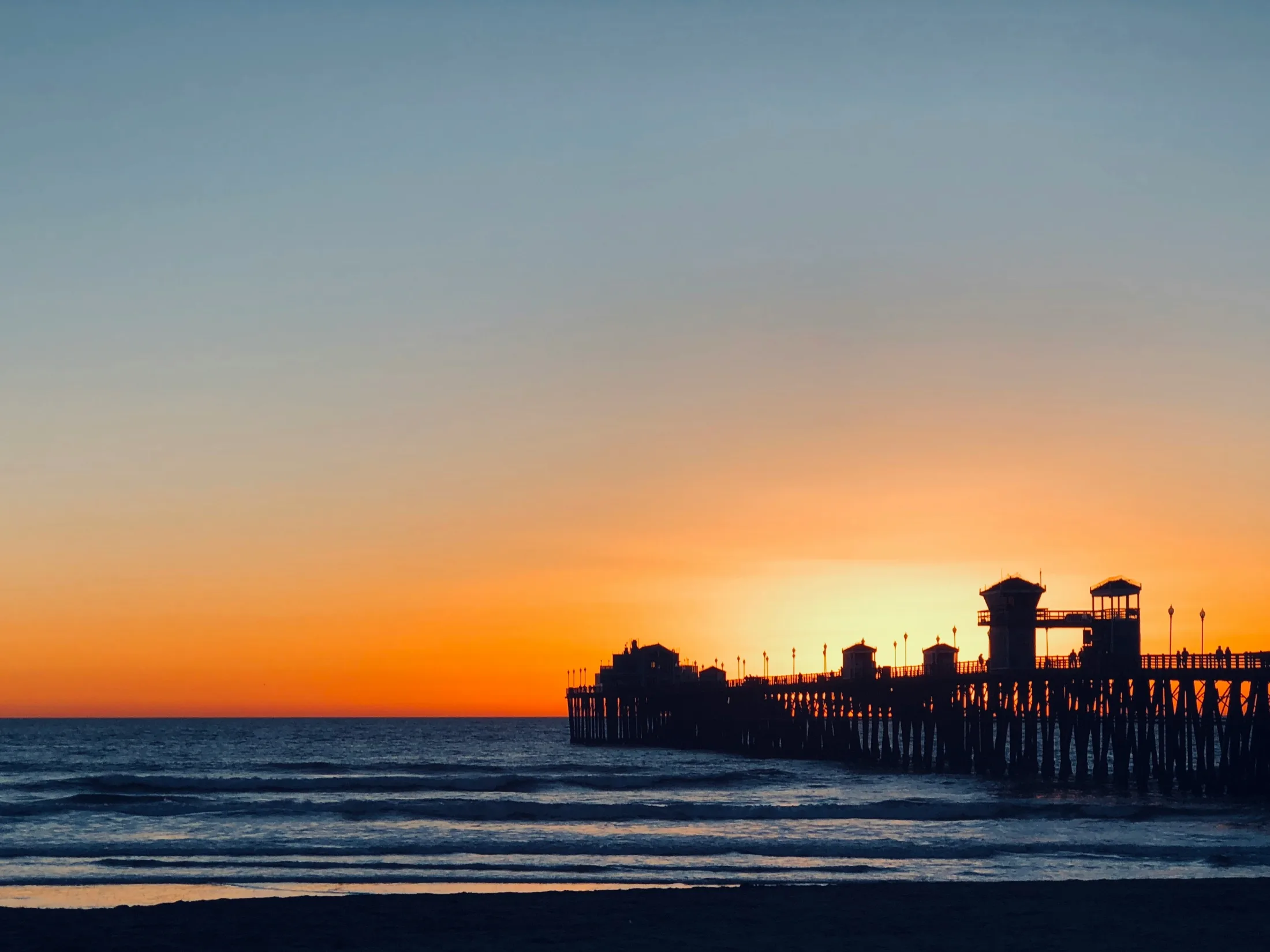Oceanside Pier silhouetted against a clean gradient sunset sky, orange fading to deep blue, calm surf in the foreground