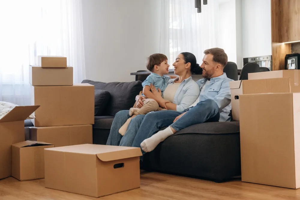 Young family — mother, father, toddler — sitting together on a couch surrounded by unpacked moving boxes, laughing