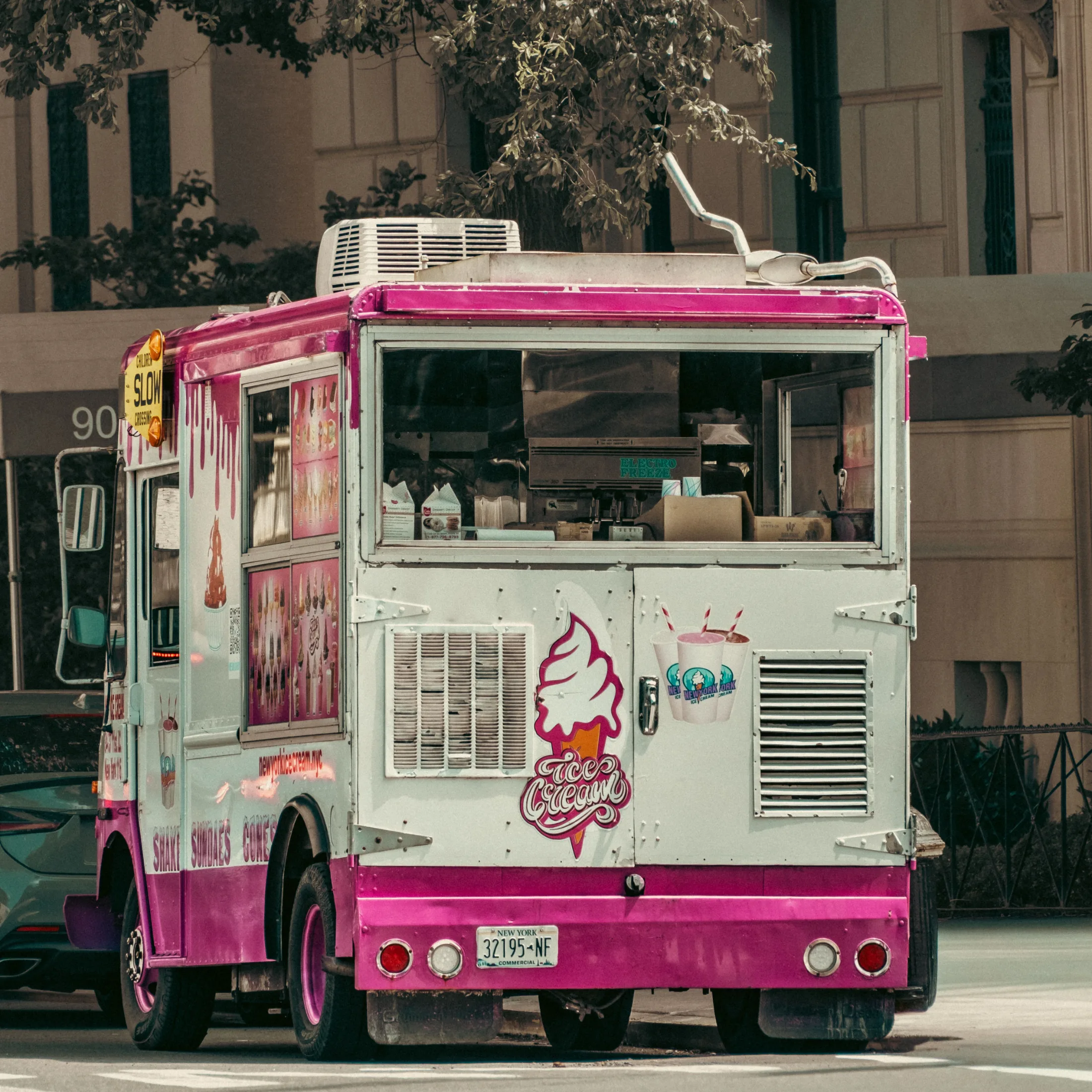A pink and white New York ice cream truck parked on a city street in summer, sundaes and cones advertised on its side panels