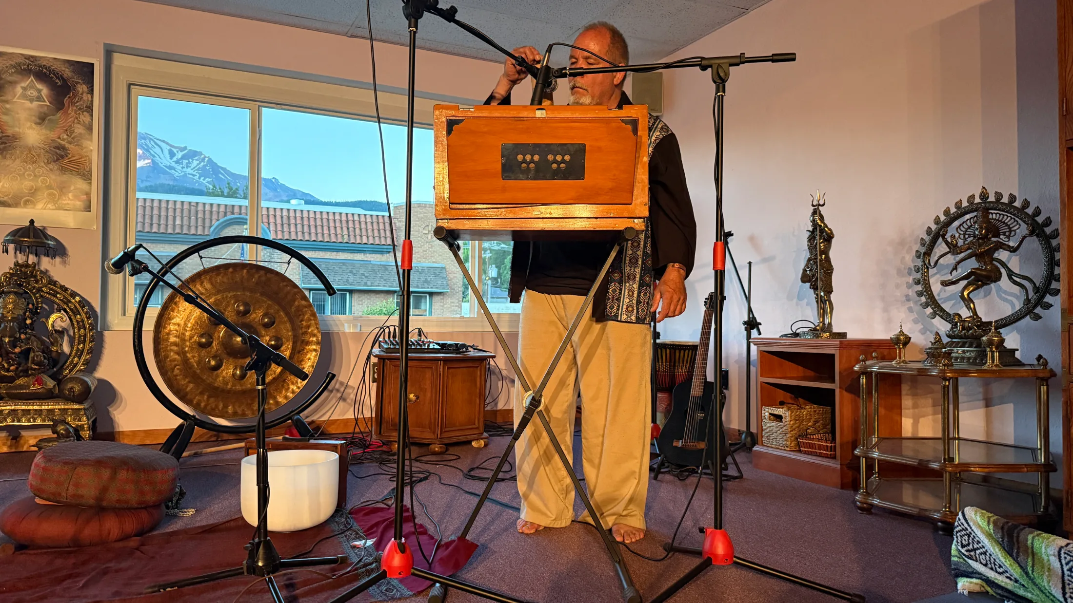 Matthew Kocel standing barefoot at a harmonium, performing at Soul Connections Community Center with Mount Shasta visible through the window behind him