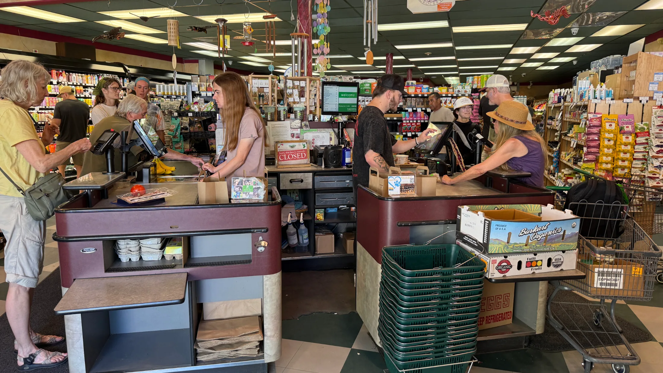 Busy checkout lanes inside Berryvale Grocery with shoppers and staff, wind chimes hanging from the ceiling
