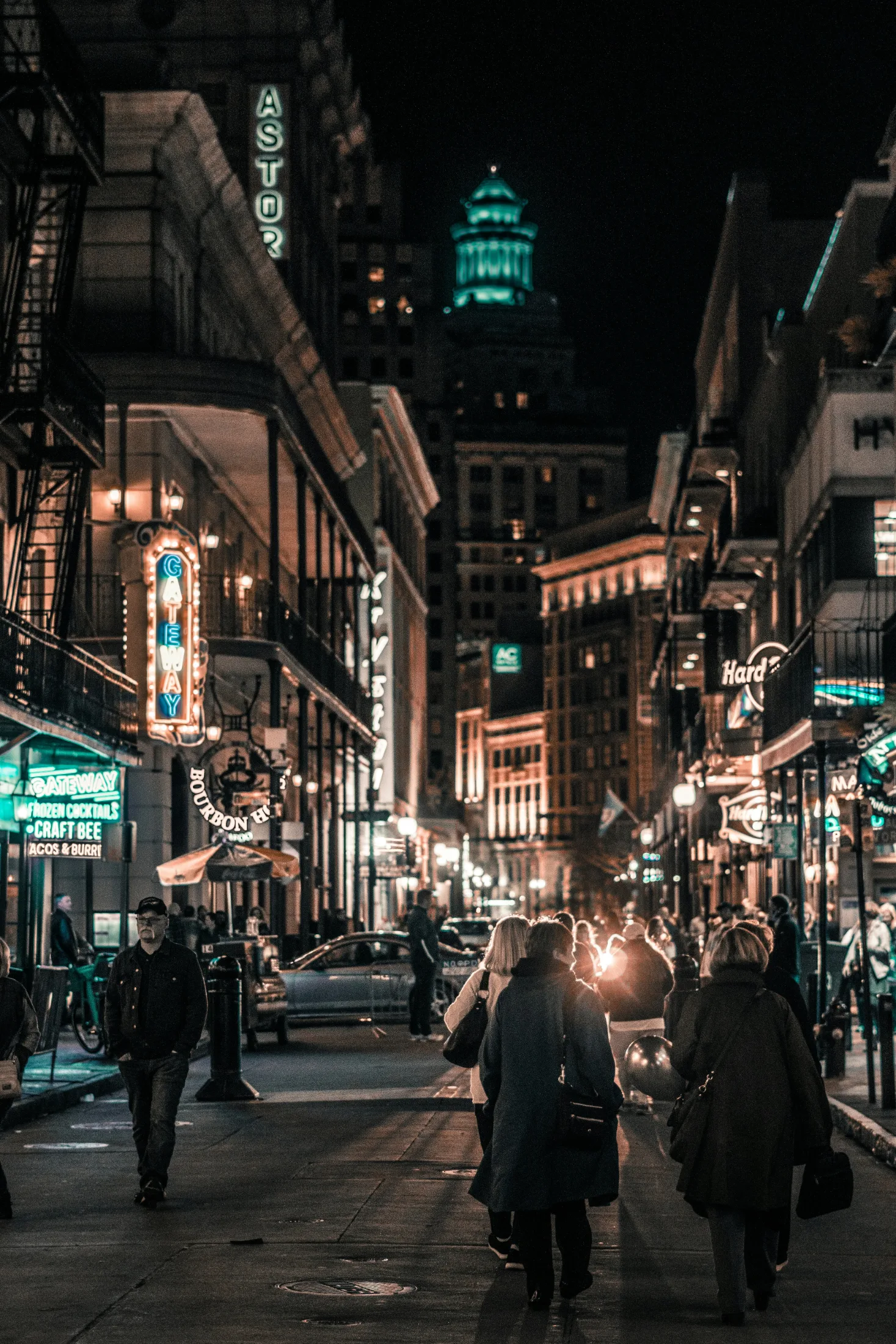A dense nighttime crowd walking a narrow New Orleans street, illuminated bar and theatre signs overhead and a green-lit tower rising in the distance.