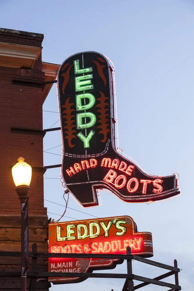 Neon boot sign for Leddy's Handmade Boots and Saddlery glowing green and red against a blue evening sky at the corner of N. Main and Exchange
