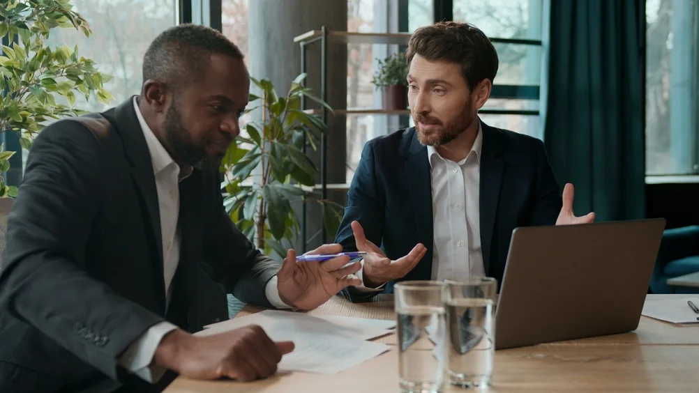 Two men in a professional setting, reviewing a laptop together — an earnest, focused conversation