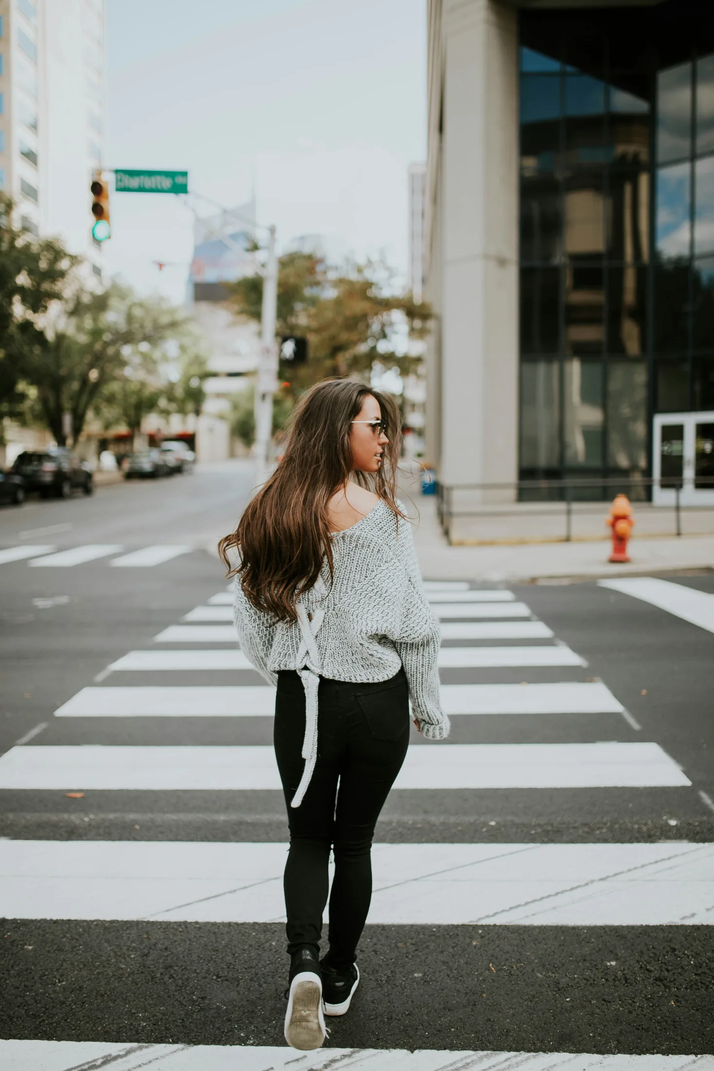 A young woman mid-stride crossing a wide city crosswalk, hair swept back, sunglasses on, buildings reflected in glass behind her