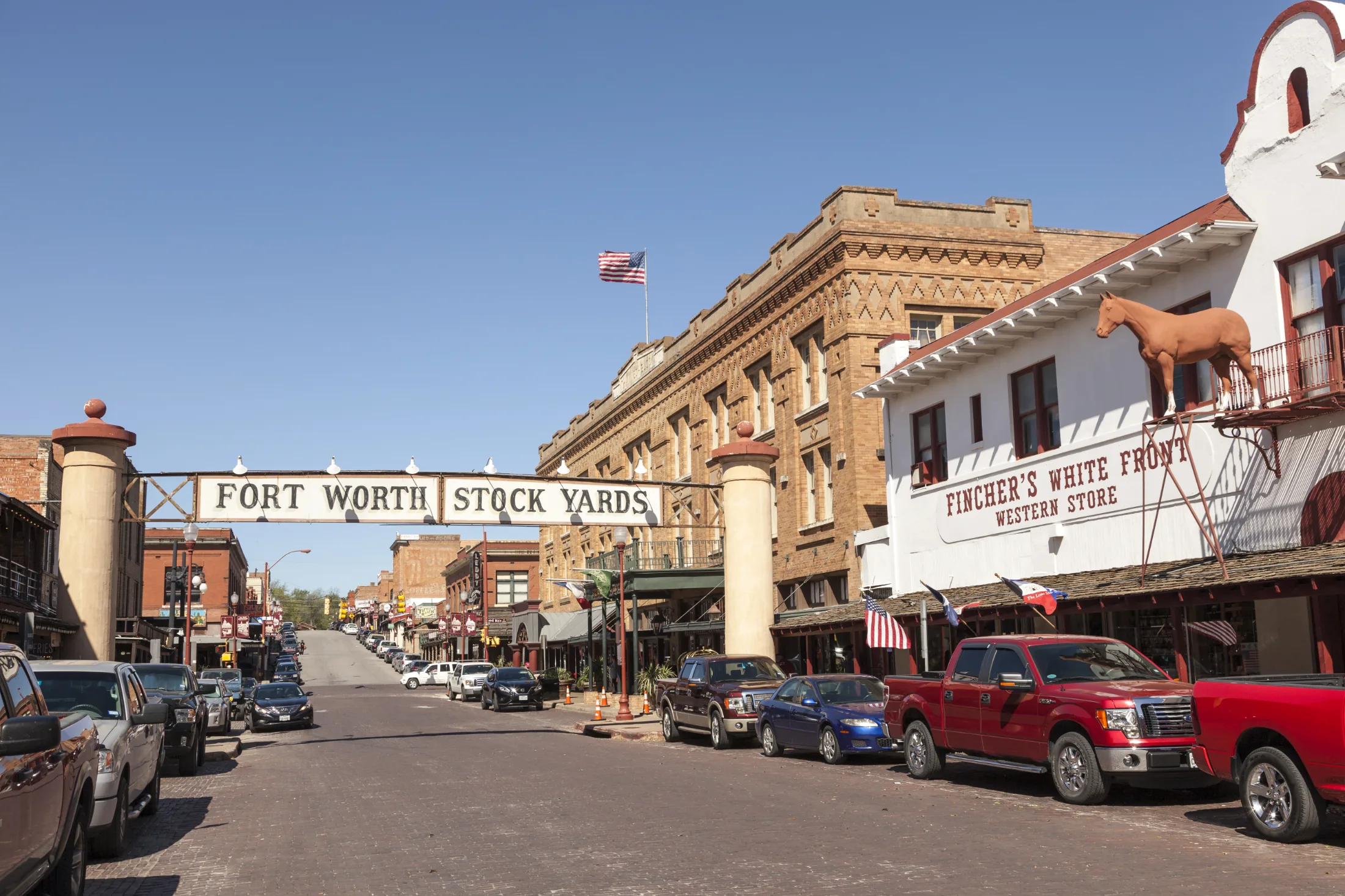 The Fort Worth Stock Yards arch spanning Exchange Avenue with Fincher's White Front Western Store and pickup trucks lining the wide brick street under a blue Texas sky