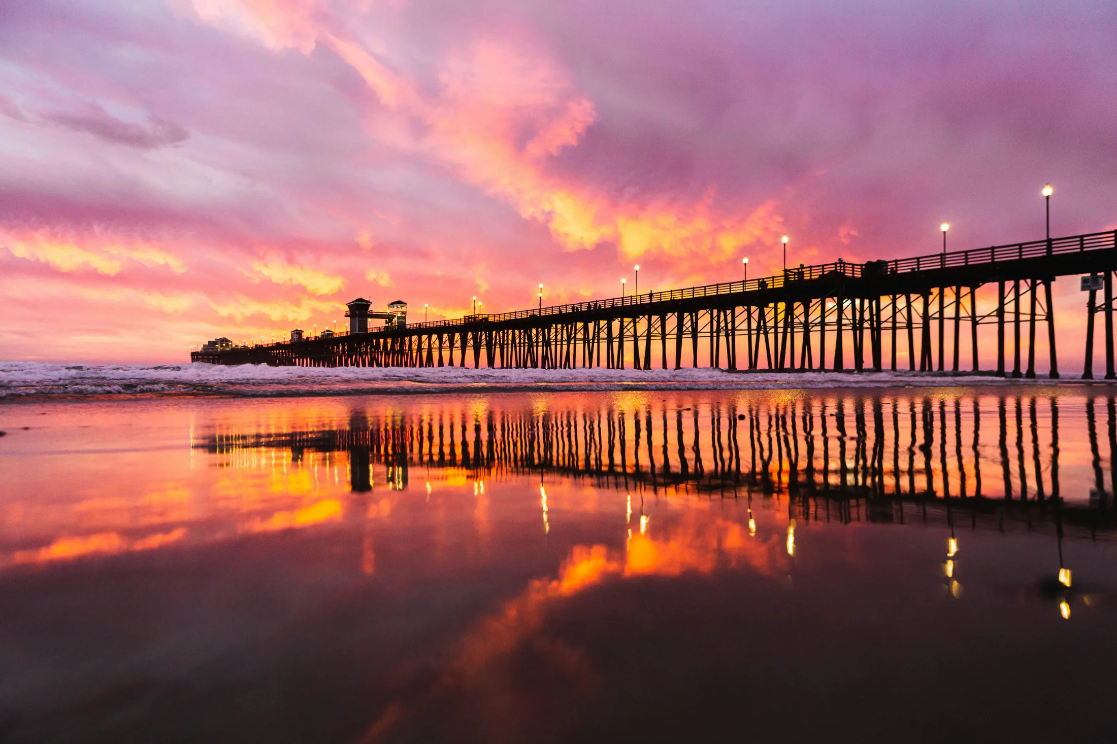 Oceanside Pier at dramatic magenta and orange sunset, full pier reflection mirrored perfectly in wet beach sand, pier lights glowing along the railing
