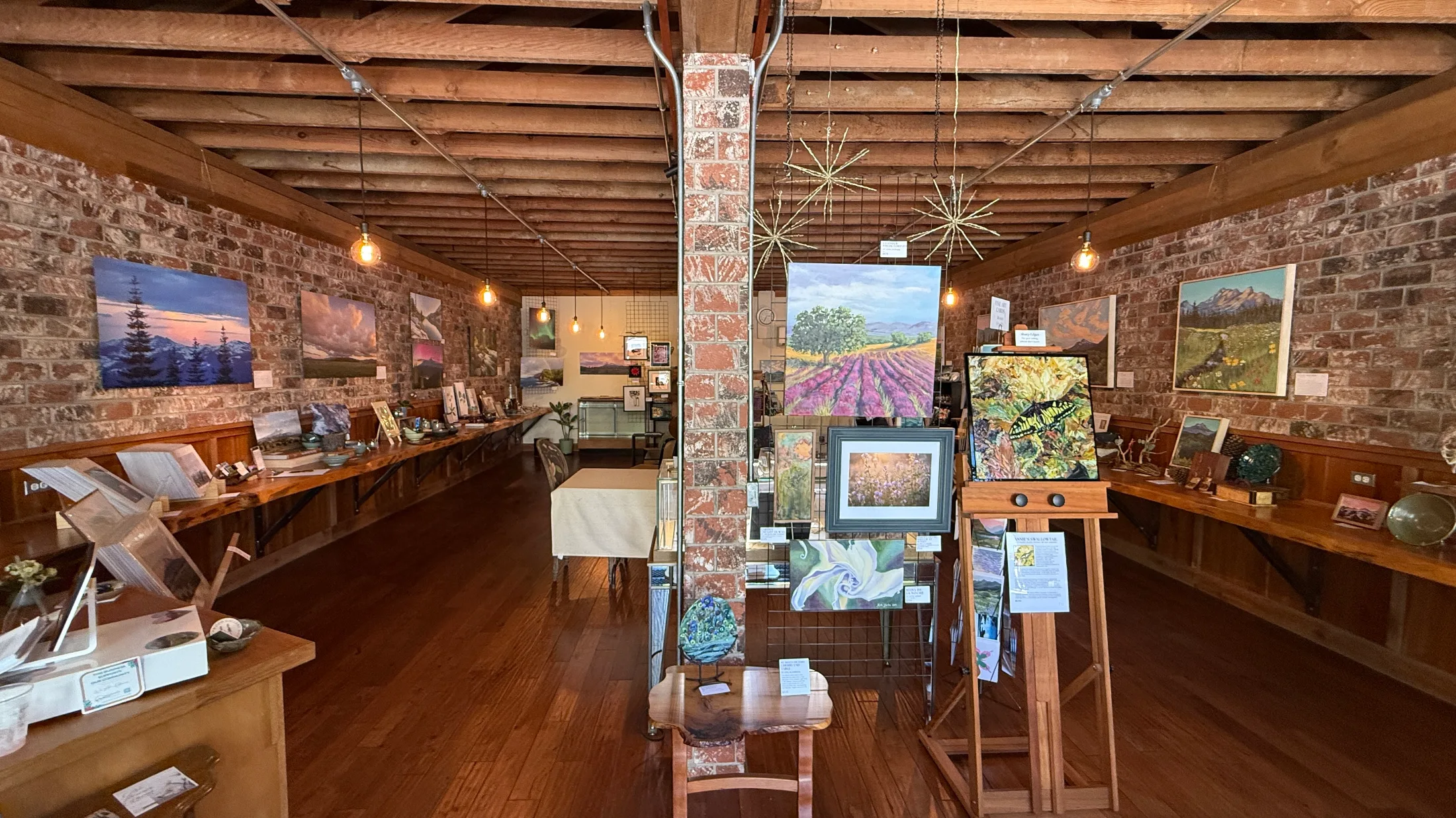 Wide view of Redwood Gallery interior showing paintings on brick walls, wooden display tables, and handmade goods