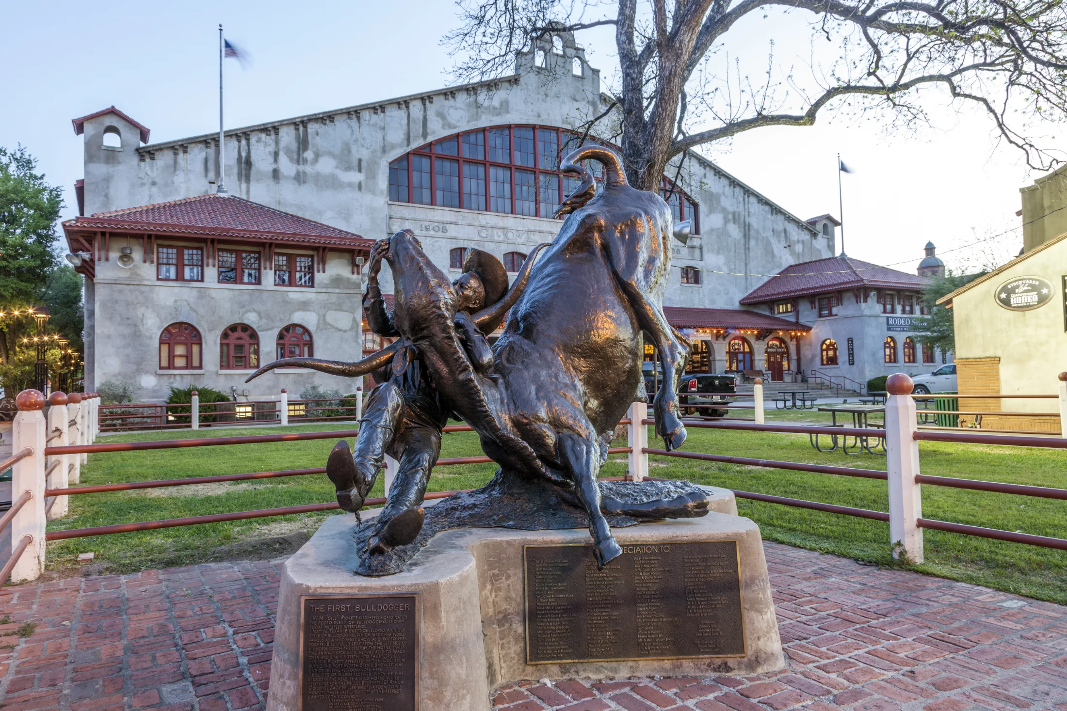 Bronze bulldogger statue mid-wrestle with a steer in front of the 1908 Coliseum at the Fort Worth Stockyards at blue hour