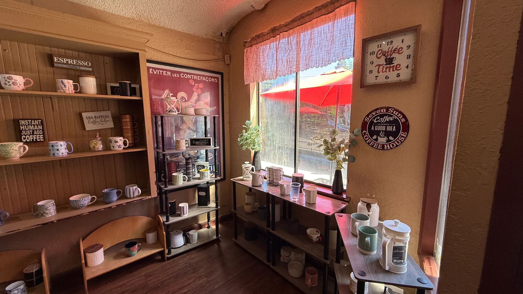 Interior nook at Seven Suns with shelves of mugs for sale, a coffee-themed clock, and a view of the red patio umbrellas through the window