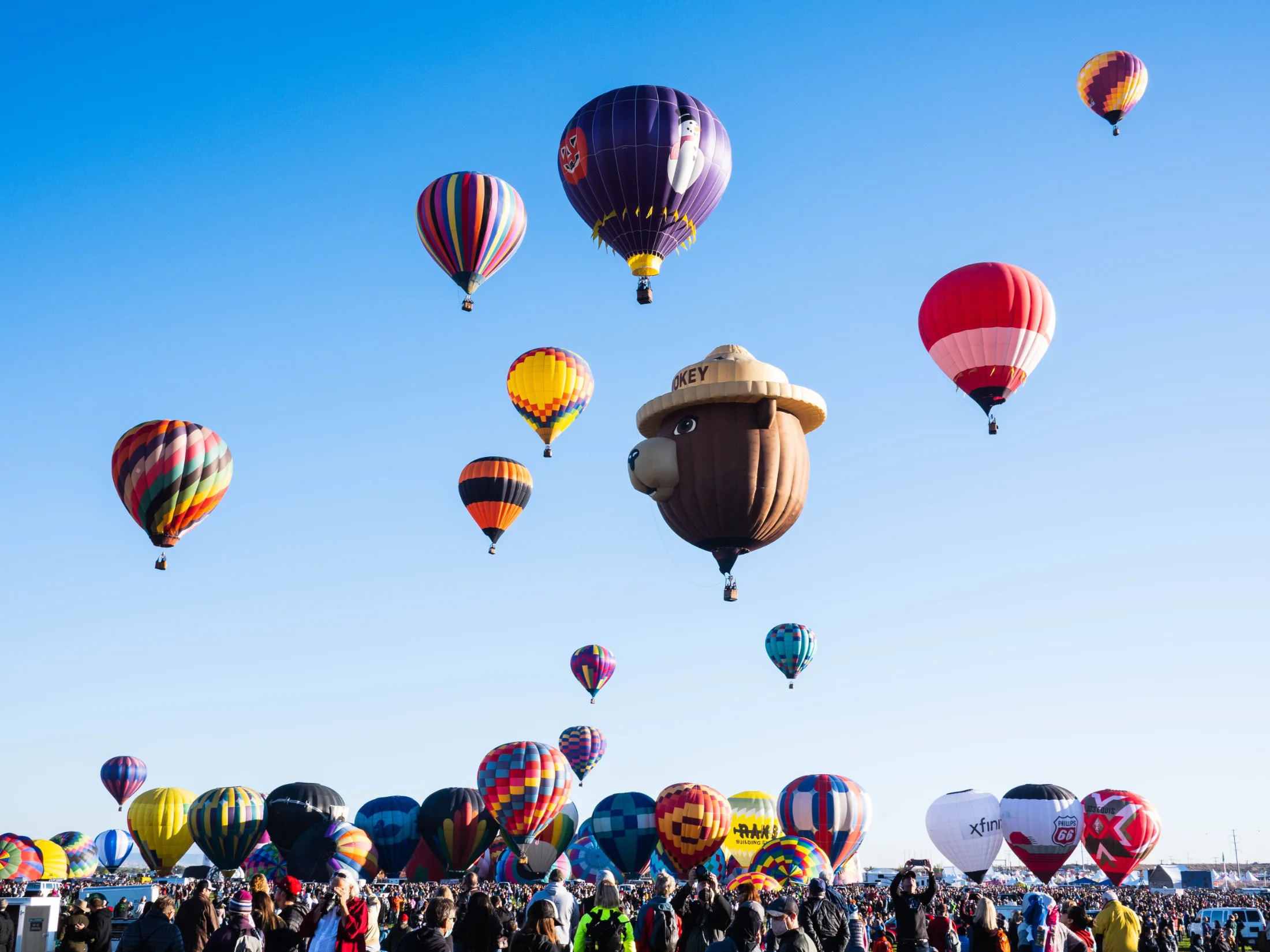 Smokey Bear novelty balloon and dozens of others rising above a dense crowd on the Balloon Fiesta launch field