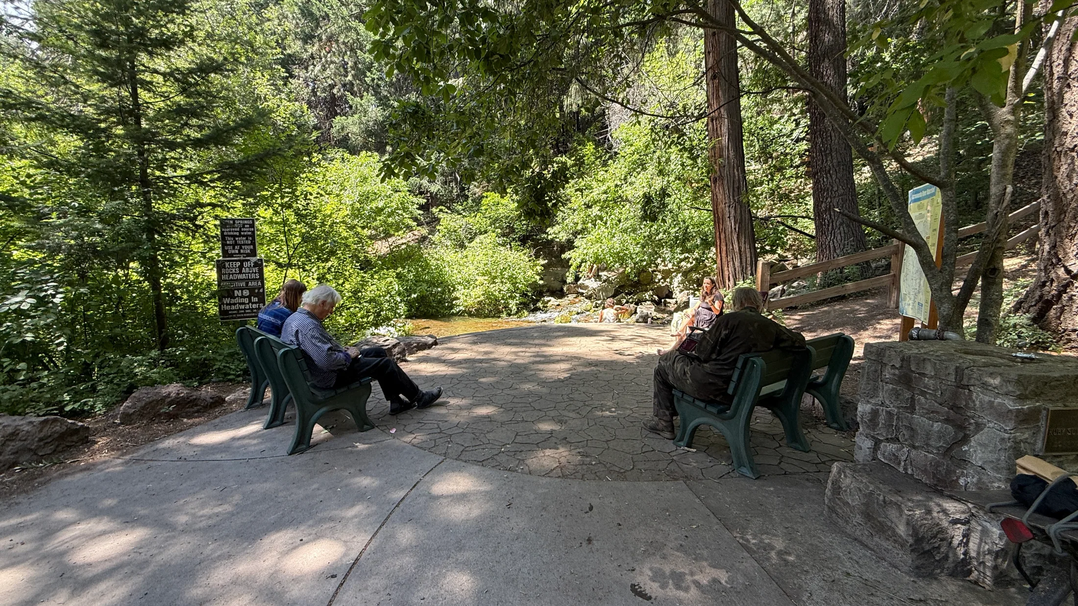 The viewing patio at Headwaters Spring with green park benches, local visitors seated, a footbridge visible to the right, and interpretive signs posted near the water