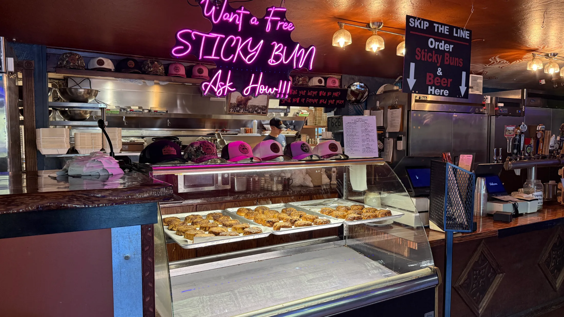 Pink neon sign reading 'Want a Free Sticky Bun? Ask How!!!' above a glass display case full of fresh sticky buns at Yaks on the 5