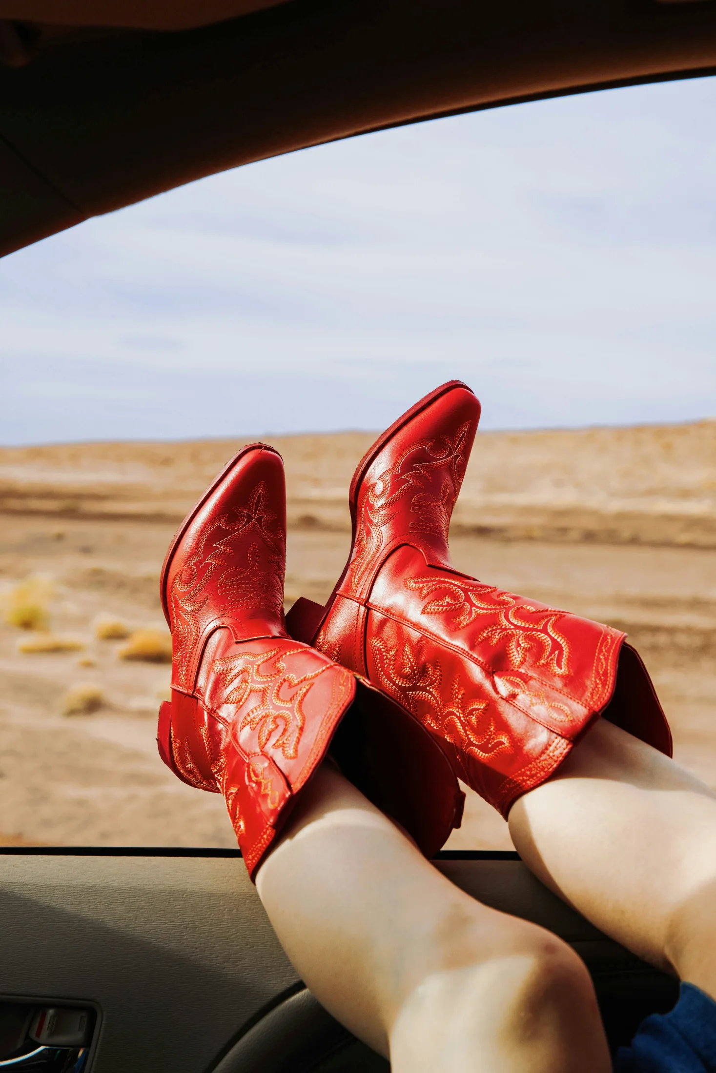 Bright red cowboy boots with gold stitching propped up on a car dashboard, wide desert scrubland stretching out the window behind them